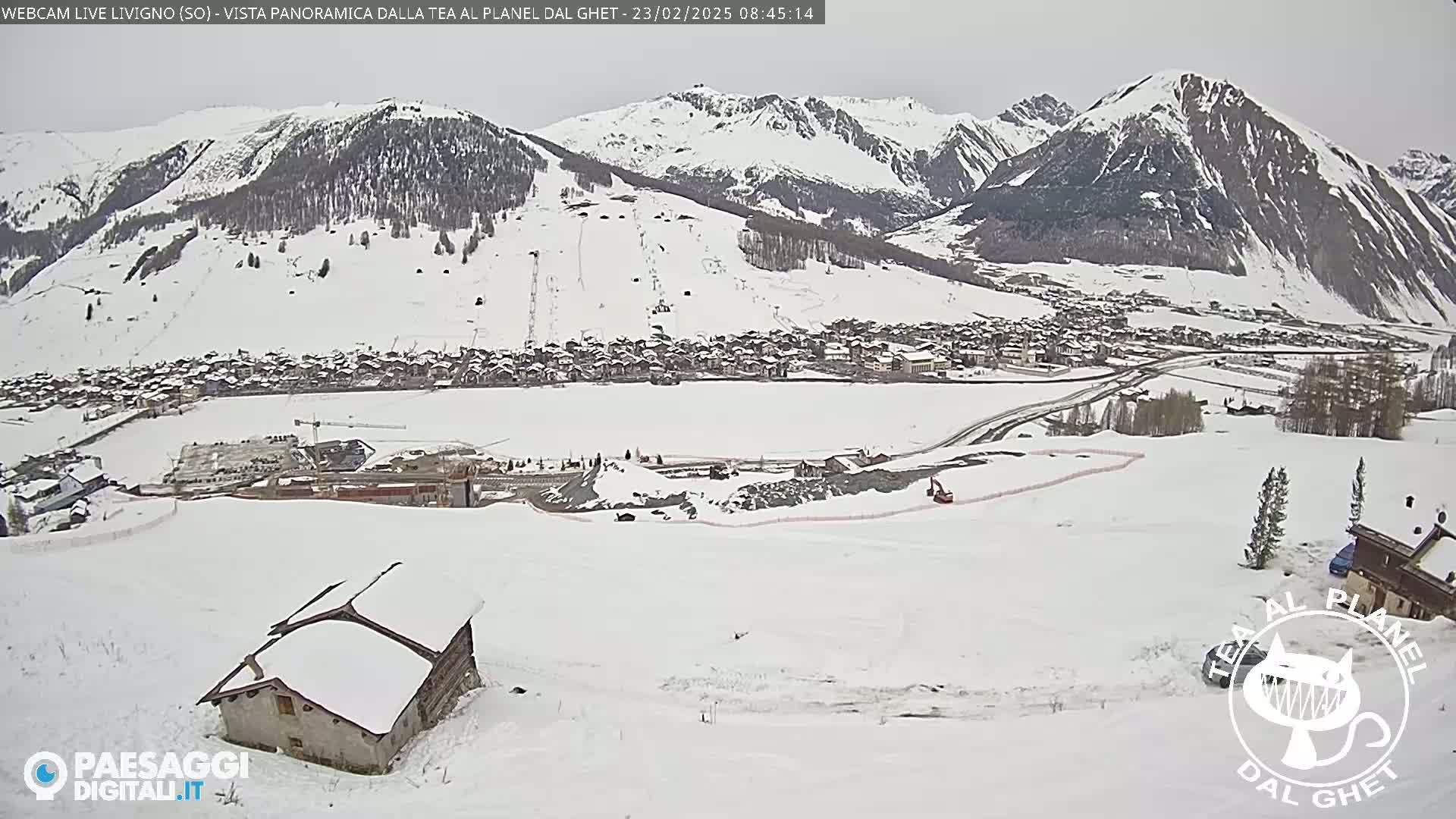 A snow-covered village nestled in a valley between snow-capped mountains, with a small snow-covered house in the foreground and construction visible in the mid-ground.