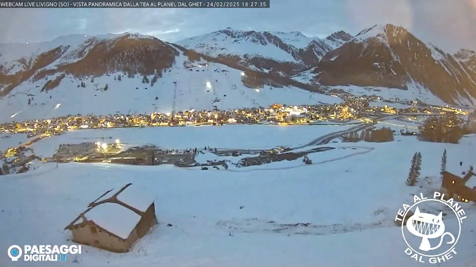 A snow-covered mountain town is illuminated at night, with a ski slope visible on the mountainside under a twilight sky.