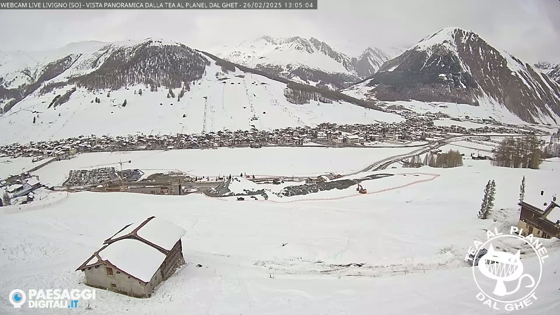 A snow-covered mountain village is viewed from a hillside, with a small snow-covered house in the foreground and overcast skies.