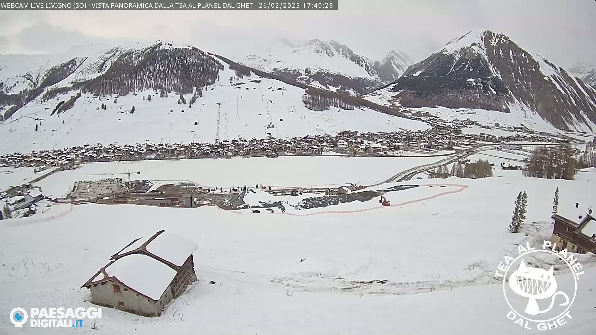 A snow-covered mountain village is seen from a high vantage point on a cloudy day, with construction visible in the foreground and a small snow-covered building in the lower left.