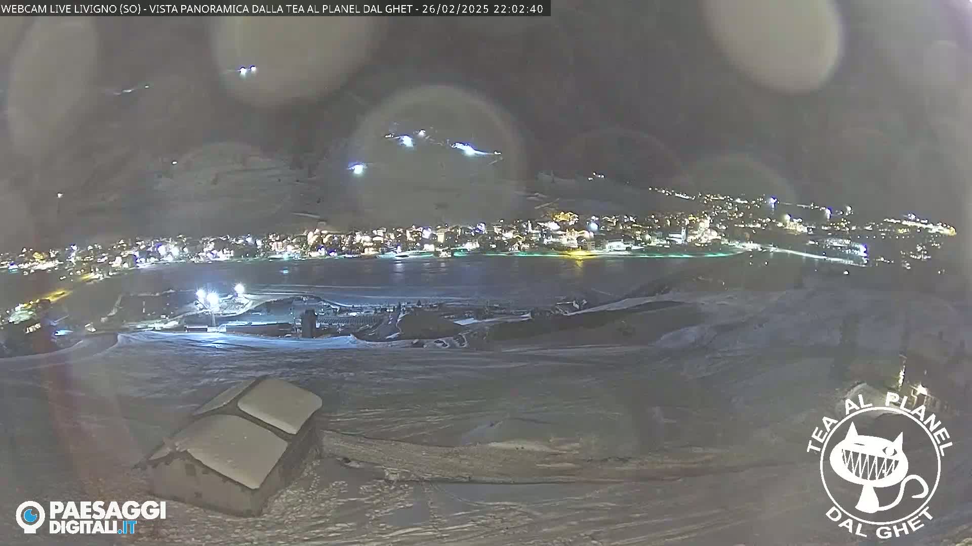 A nighttime panoramic view of a snow-covered town, illuminated by lights, under a dark, possibly slightly hazy sky.