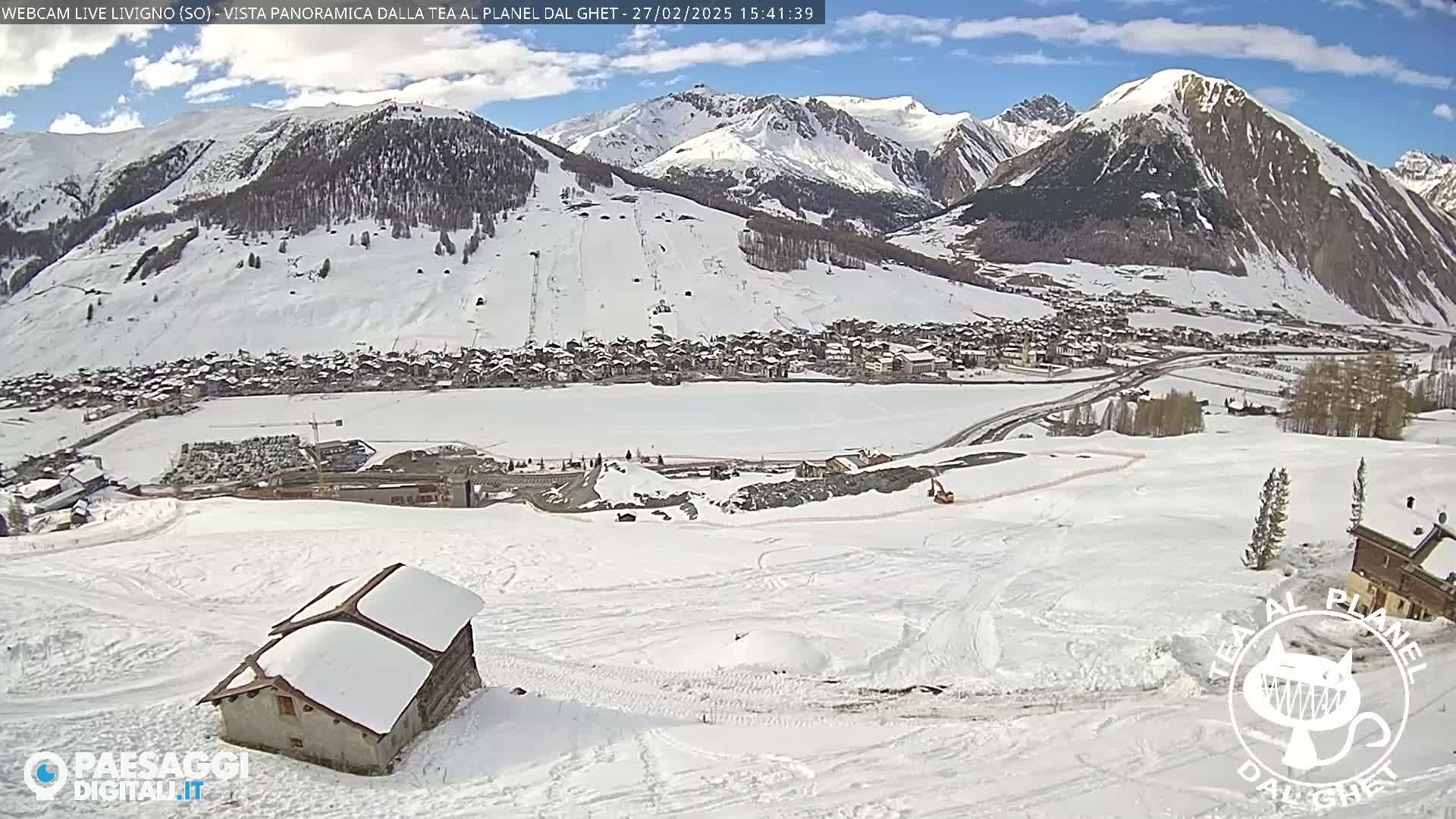 A snow-covered mountain village is seen from a high vantage point on a sunny day, with ski slopes and a small building in the foreground.