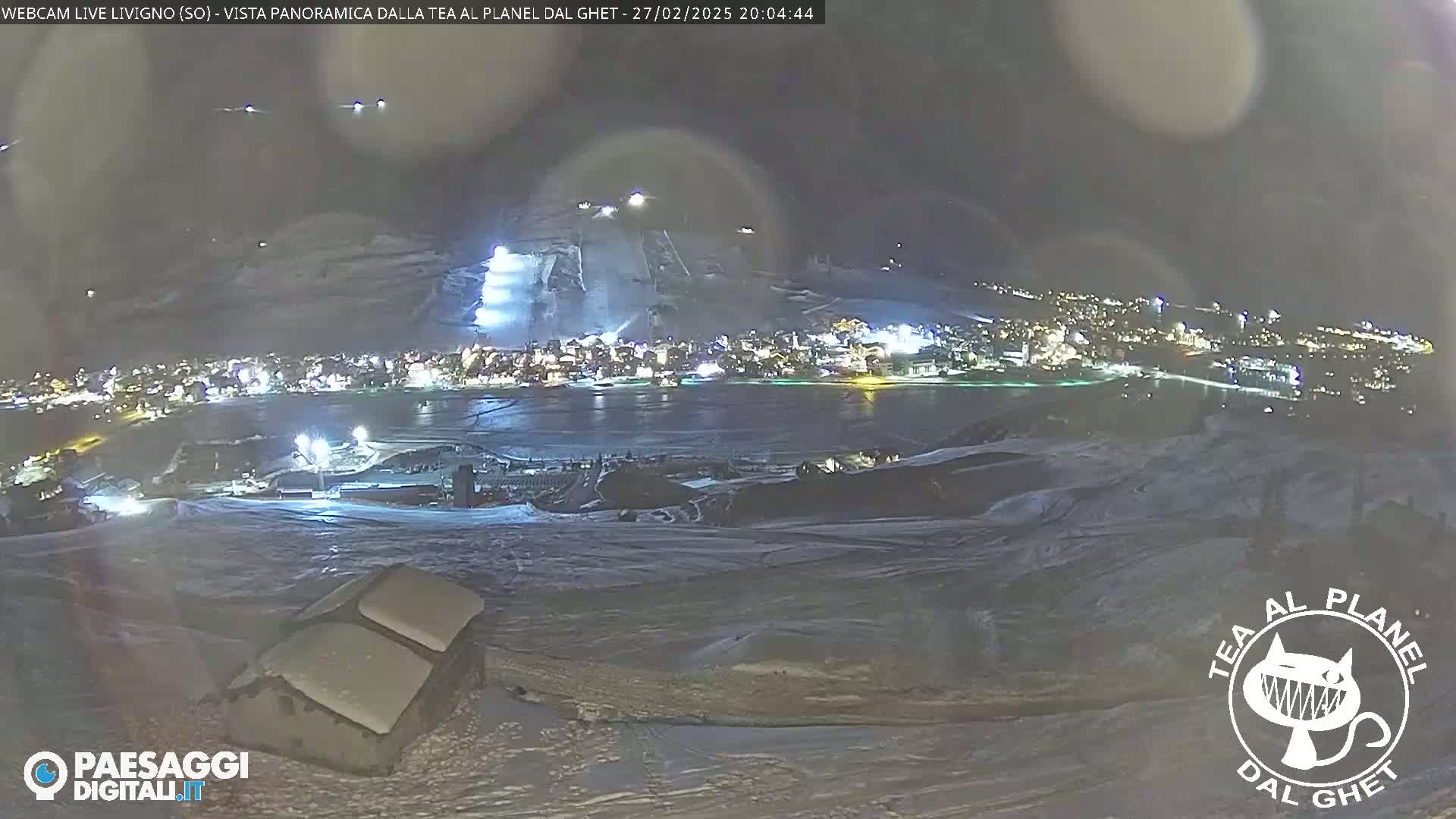 A nighttime, snowy panorama shows a brightly lit town nestled beside a dark body of water, with snow-covered slopes and a building in the foreground.