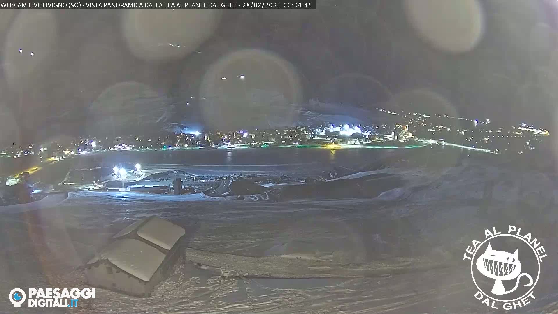 A nighttime, snowy landscape shows a town illuminated in the distance under a hazy sky.