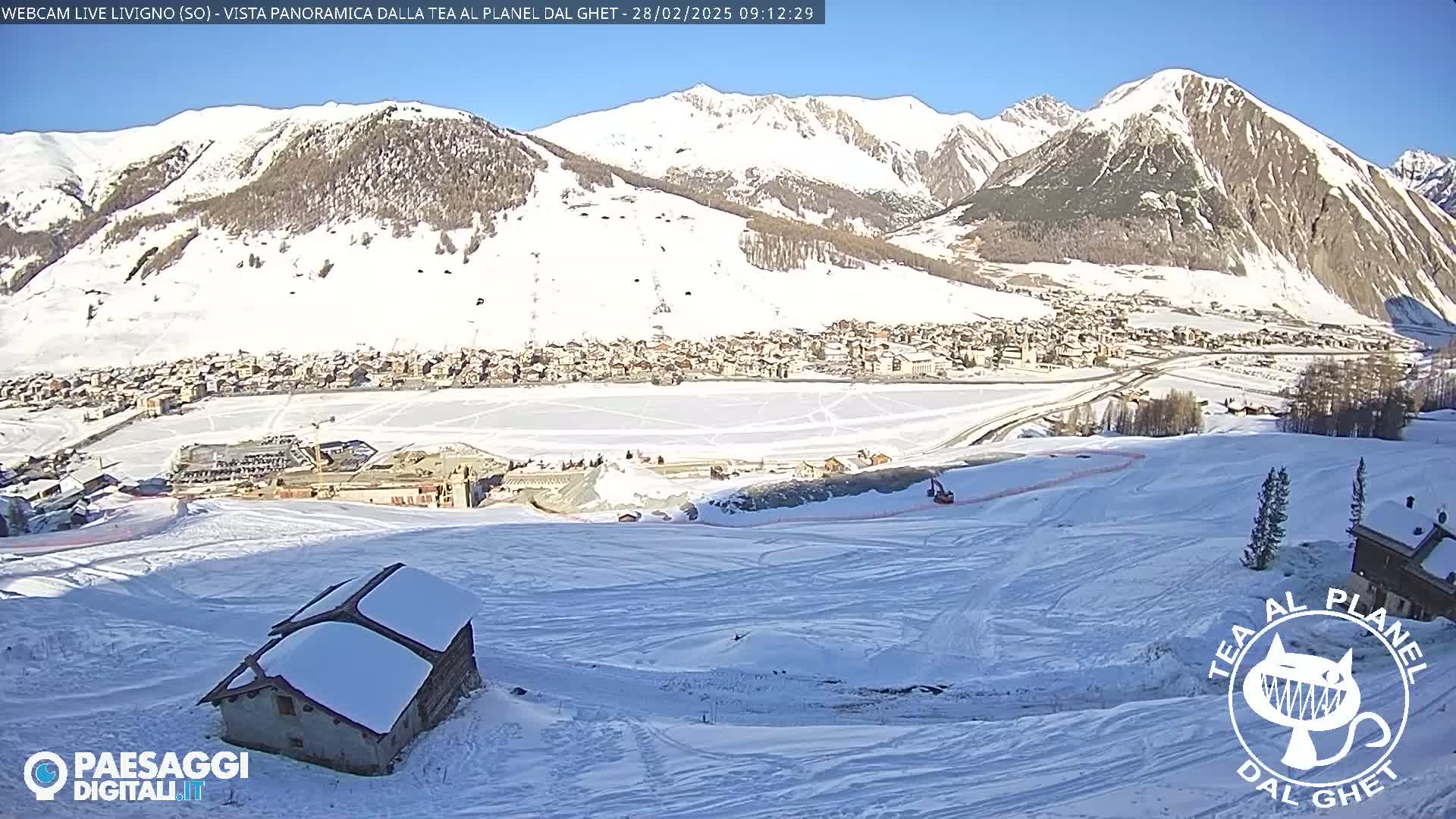 A snow-covered mountain village is seen from a high vantage point on a sunny day, with a small snow-covered building in the foreground.