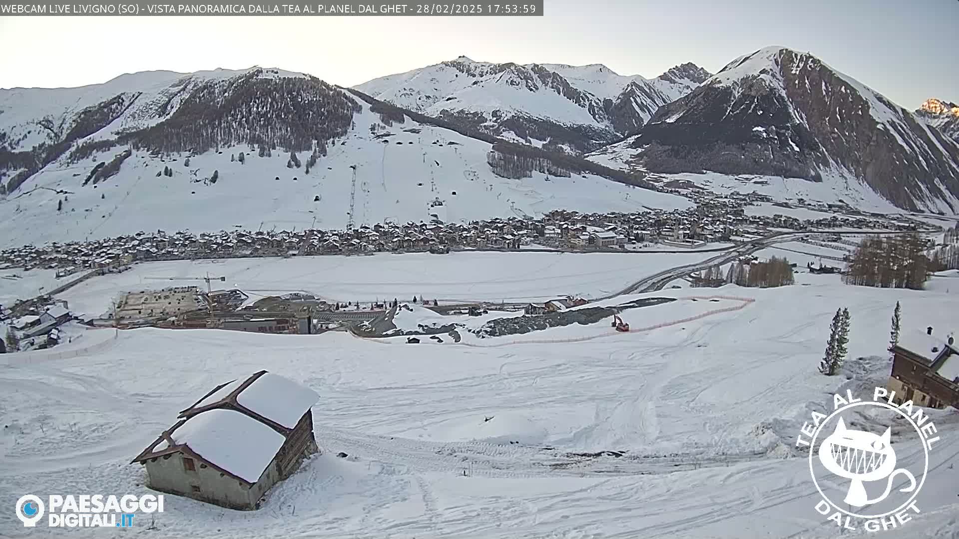 A snow-covered mountain village is seen from a snowy hillside, with mountains in the background under a clear, bright sky.