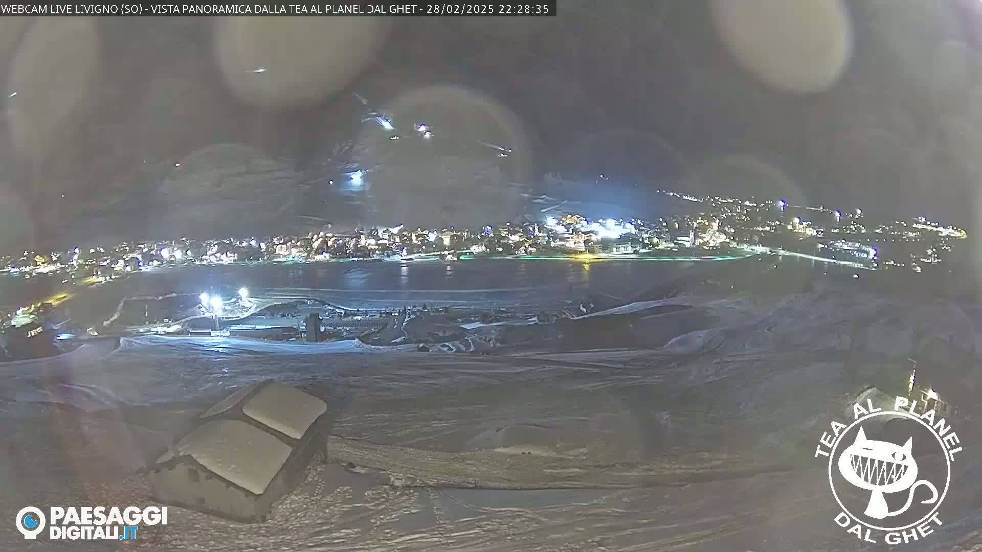 A nighttime, snowy landscape shows a town lit up in the distance, with a body of water separating the foreground from the town.