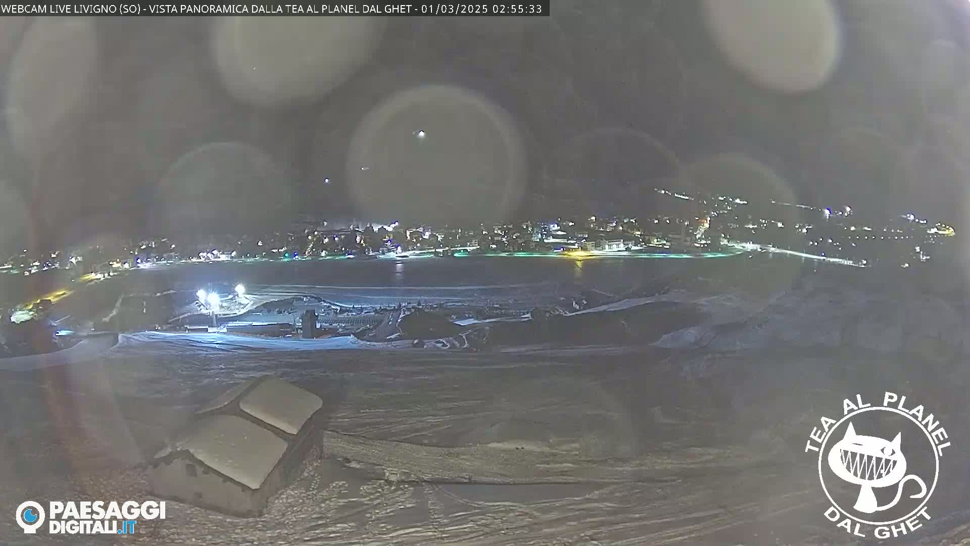 A nighttime, snowy landscape shows a town illuminated in the distance, with a structure in the foreground under a cloudy sky.