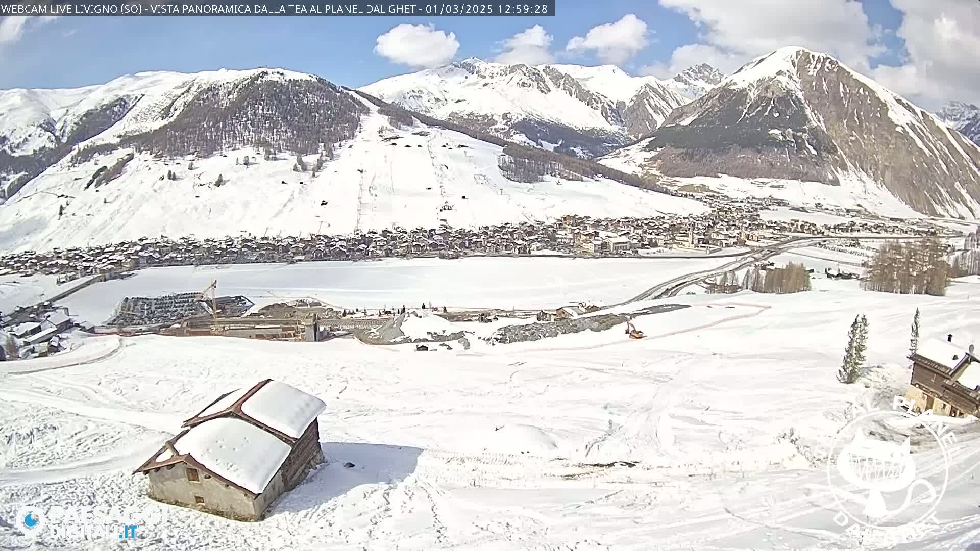 A snow-covered mountain village is seen from a high vantage point on a partly sunny day, with a small snow-covered building in the foreground and construction visible in the midground.