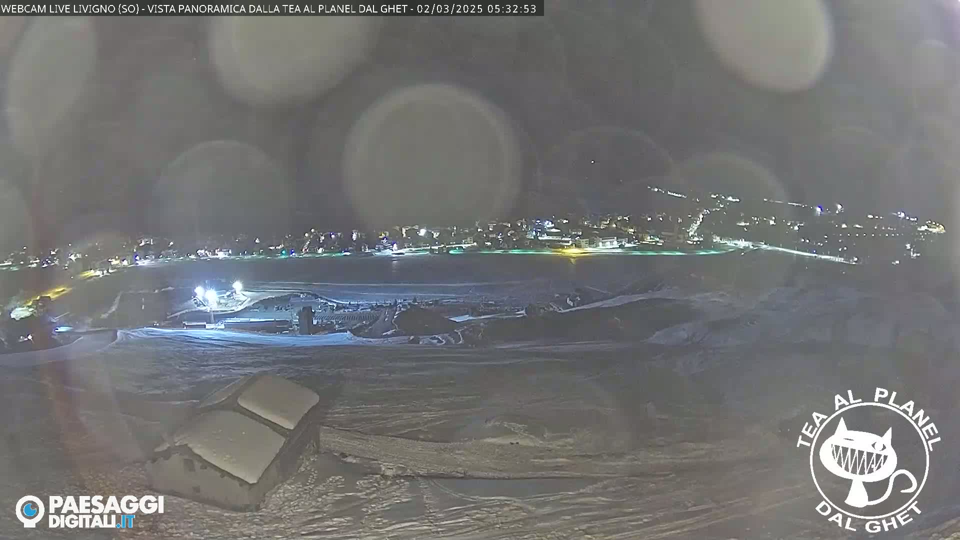 A nighttime, snowy, panoramic view of a town illuminated by lights, from a high vantage point, with some light snow falling.