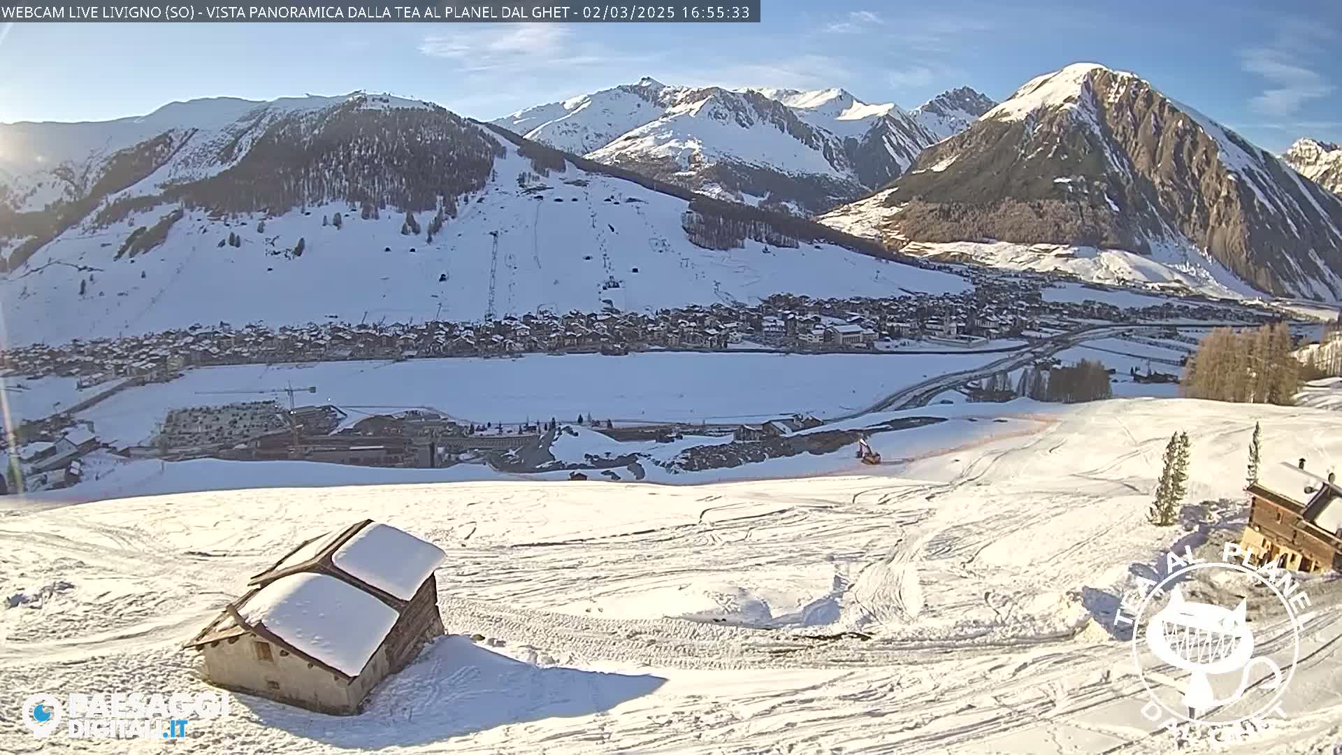 A snow-covered mountain village is visible in the distance, under a sunny sky, with a small snow-covered building in the foreground of a mostly flat, snow-covered slope showing tire tracks.