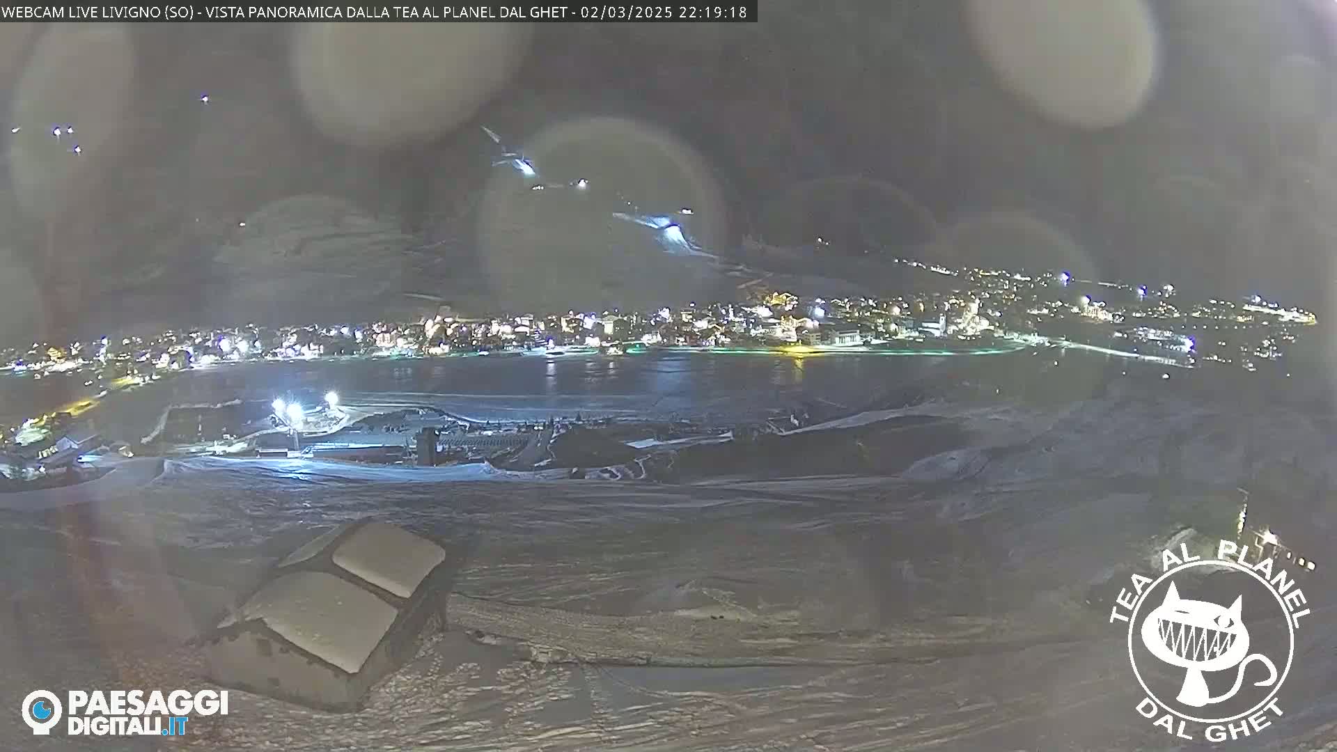 A nighttime, panoramic view of a snow-covered landscape shows a town illuminated below, with faint lights possibly from skiers or other activities on the slopes above.
