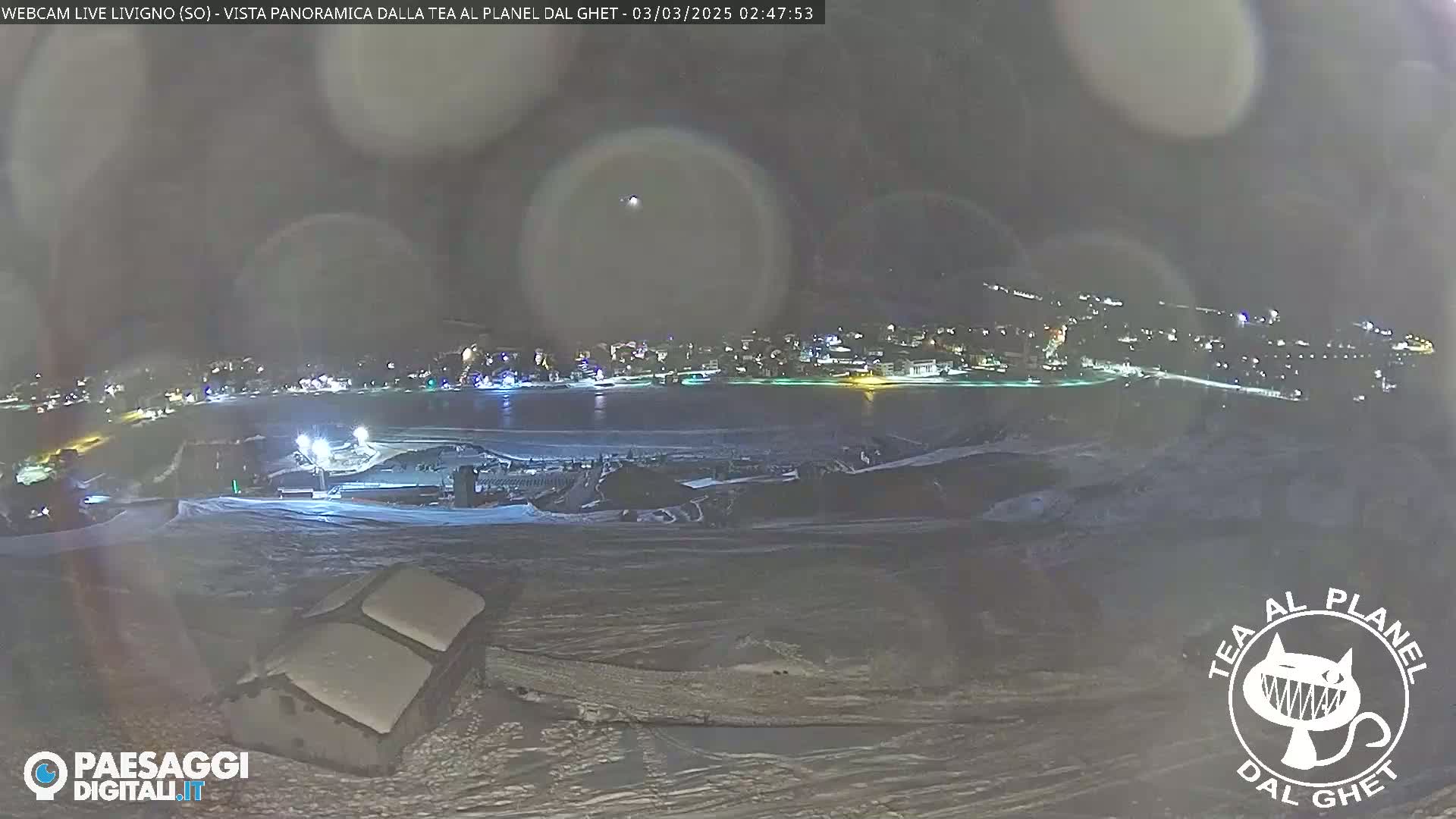A nighttime, snowy landscape shows a distant town illuminated against a dark sky, with a snow-covered foreground and a structure in the lower left corner.
