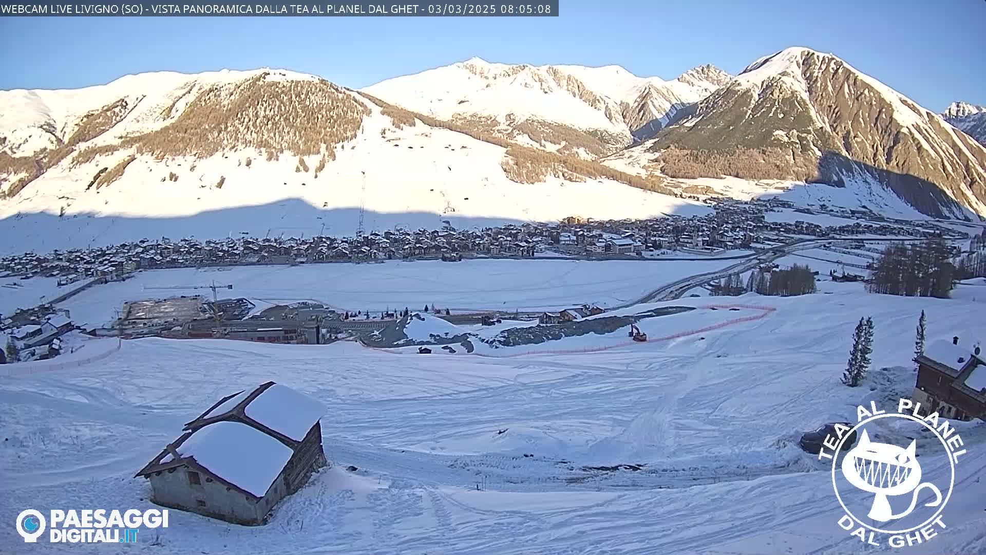 A snow-covered mountain village is nestled in a valley beneath snow-capped mountains on a sunny day.