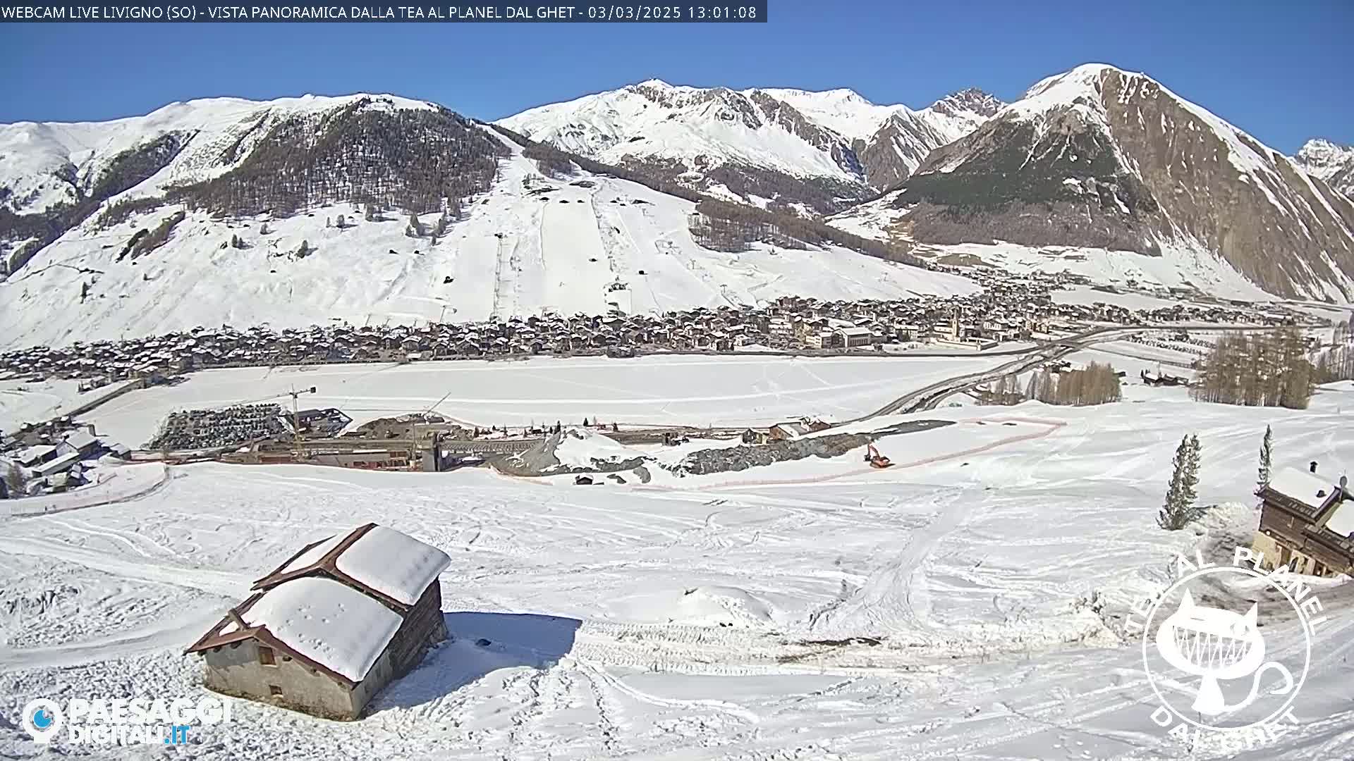 A snow-covered mountain village is visible beneath a sunny sky, with a small snow-covered house in the foreground and ski slopes on the mountainside.