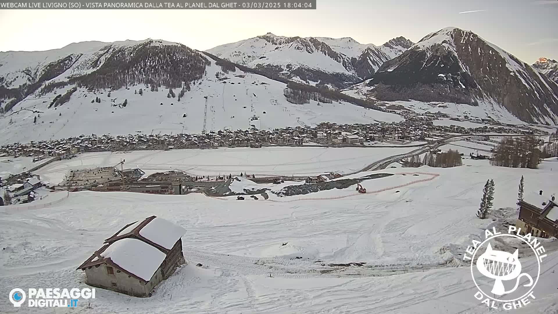 A snow-covered mountain valley town is viewed from a snowy hillside, with a small snow-covered building in the foreground.