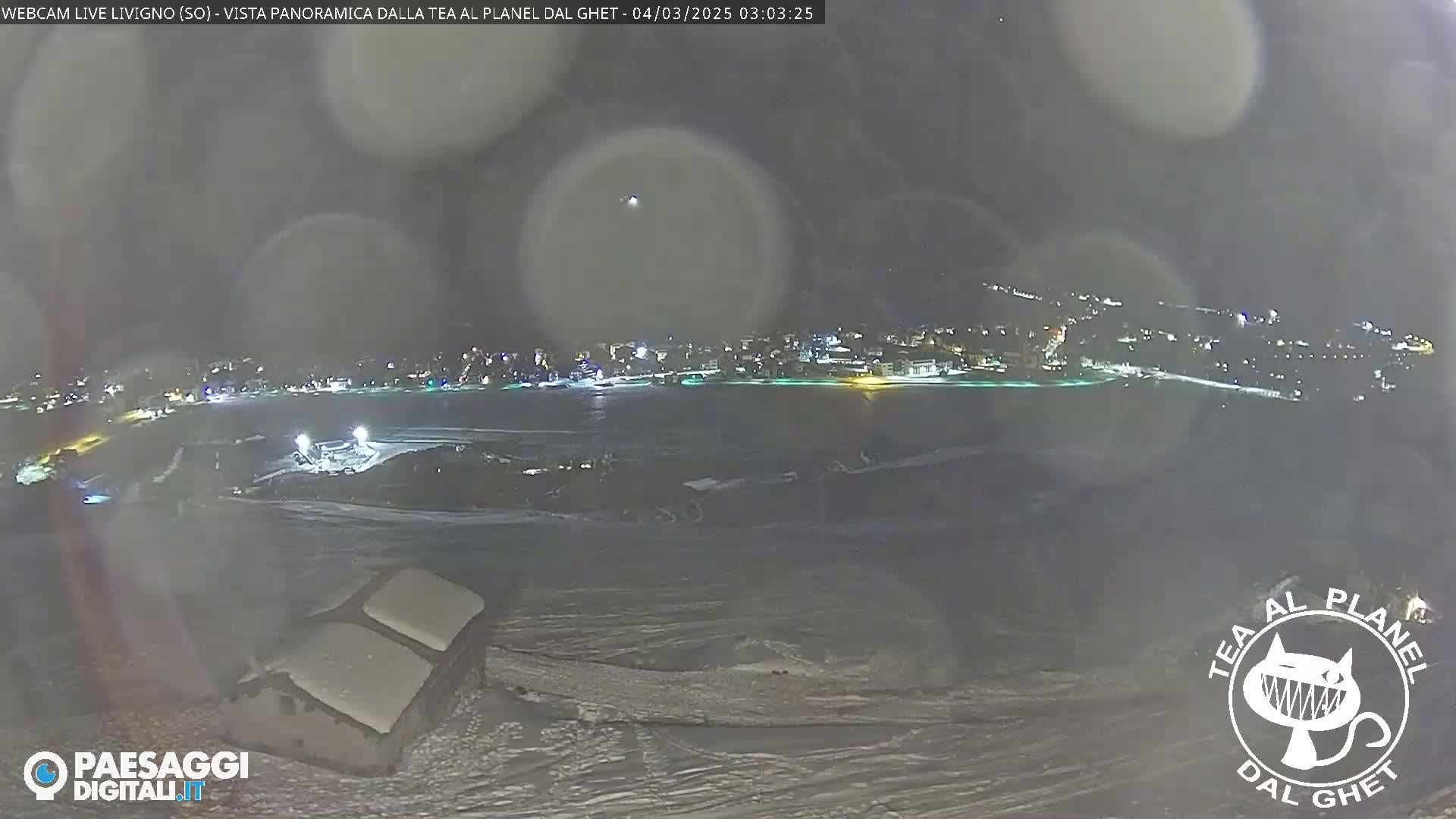 A nighttime, snowy panorama shows a town's lights across a body of water, viewed from a snow-covered vantage point.