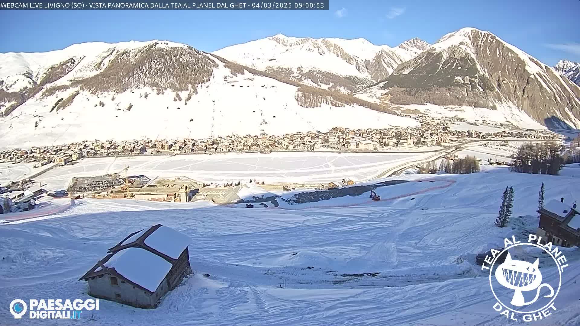 A snow-covered mountain village is nestled in a valley beneath snow-capped mountains on a sunny day.