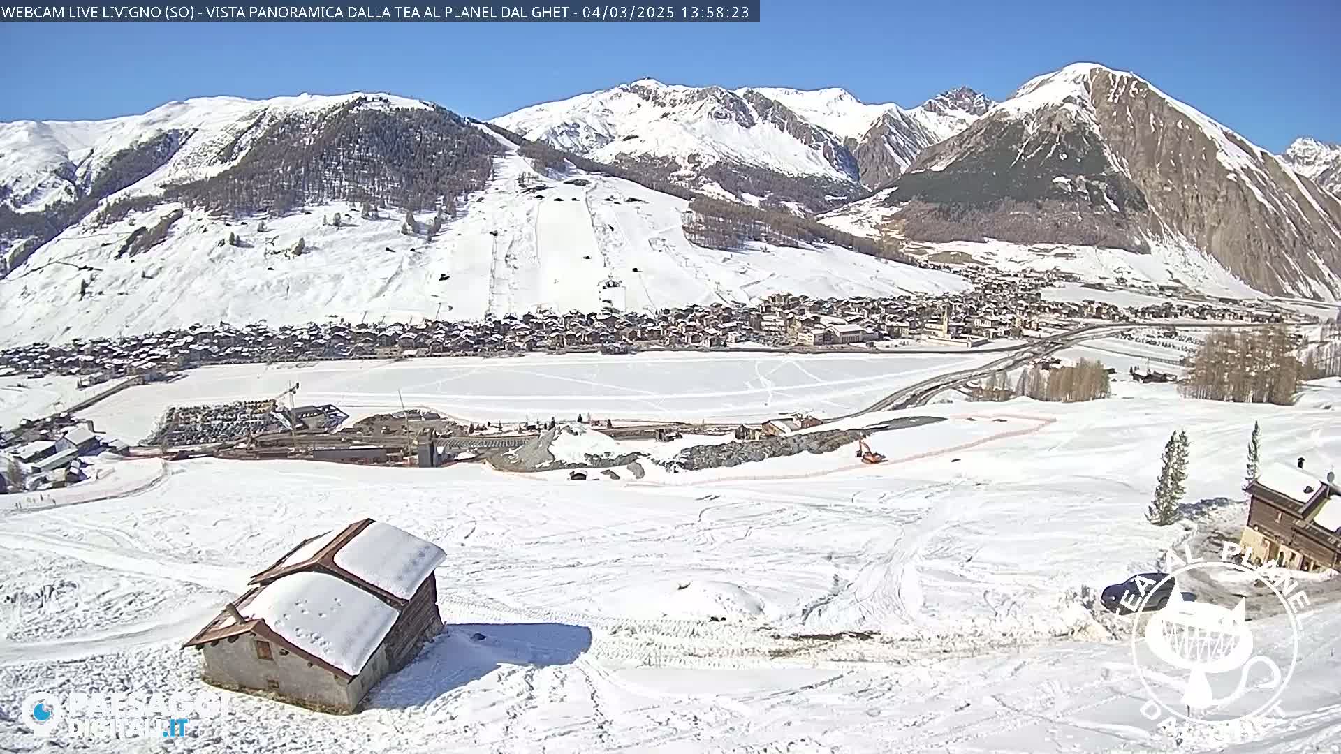 A snow-covered mountain village is seen from a high vantage point on a sunny day, with a small snow-covered building in the foreground.