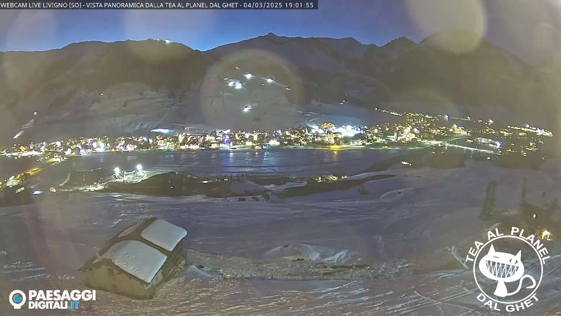 A nighttime, snowy panorama shows a town illuminated below a mountain slope with ski tracks visible under a dark sky.