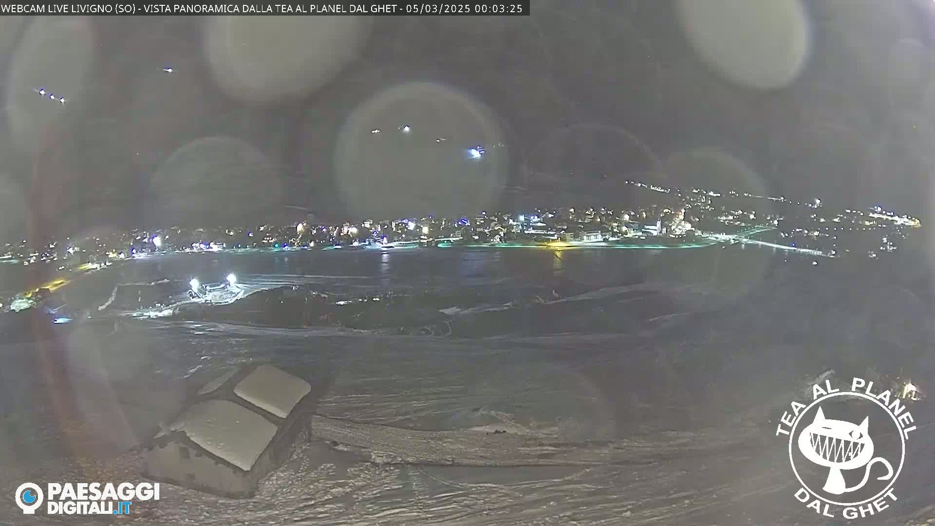 A nighttime, snowy, panoramic view of a town's lights across a body of water, with several small, distant lights possibly aircraft in the sky.