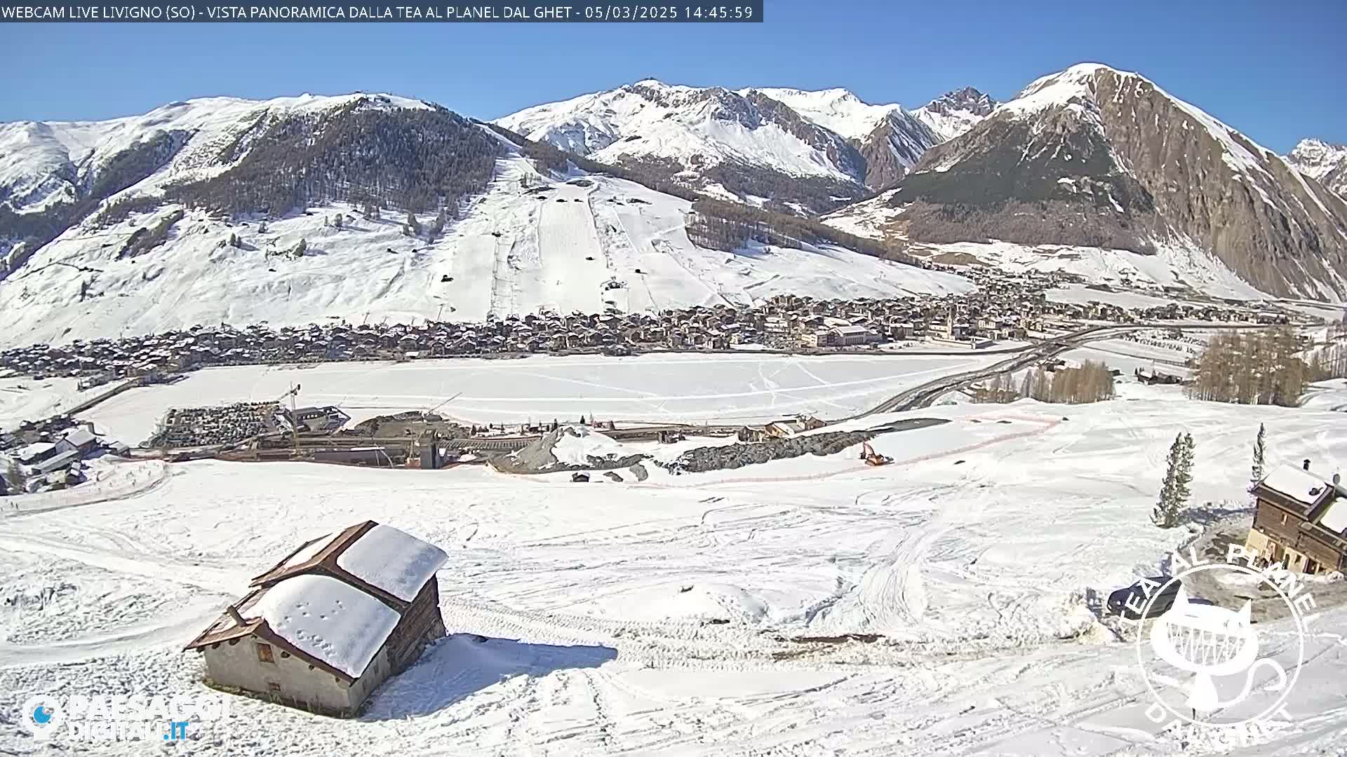 A snow-covered mountain valley, under a sunny sky, shows a village nestled below ski slopes and a small building in the foreground.