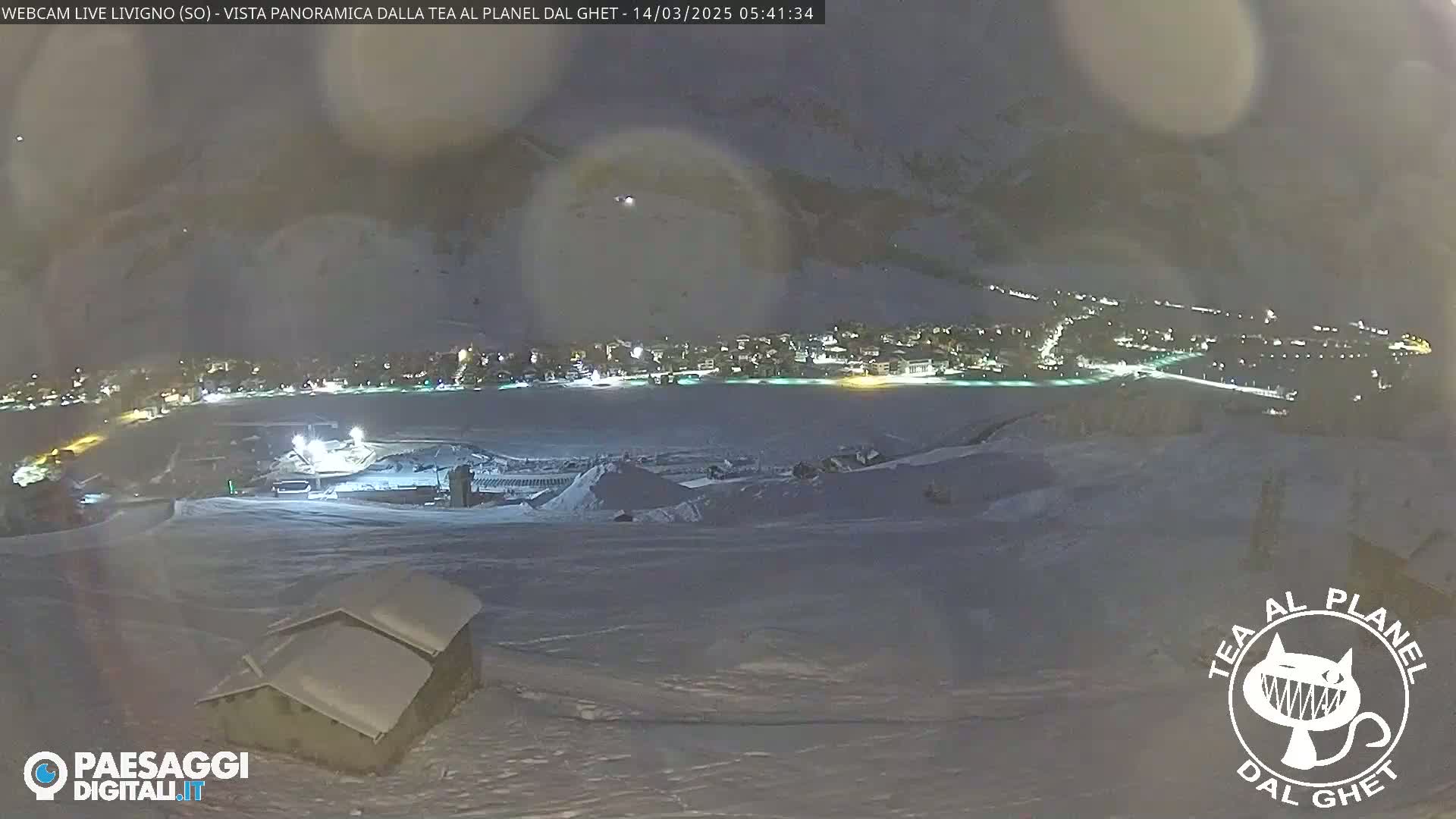 A nighttime, snowy panorama shows a village nestled in a valley, illuminated by lights, under a hazy sky.