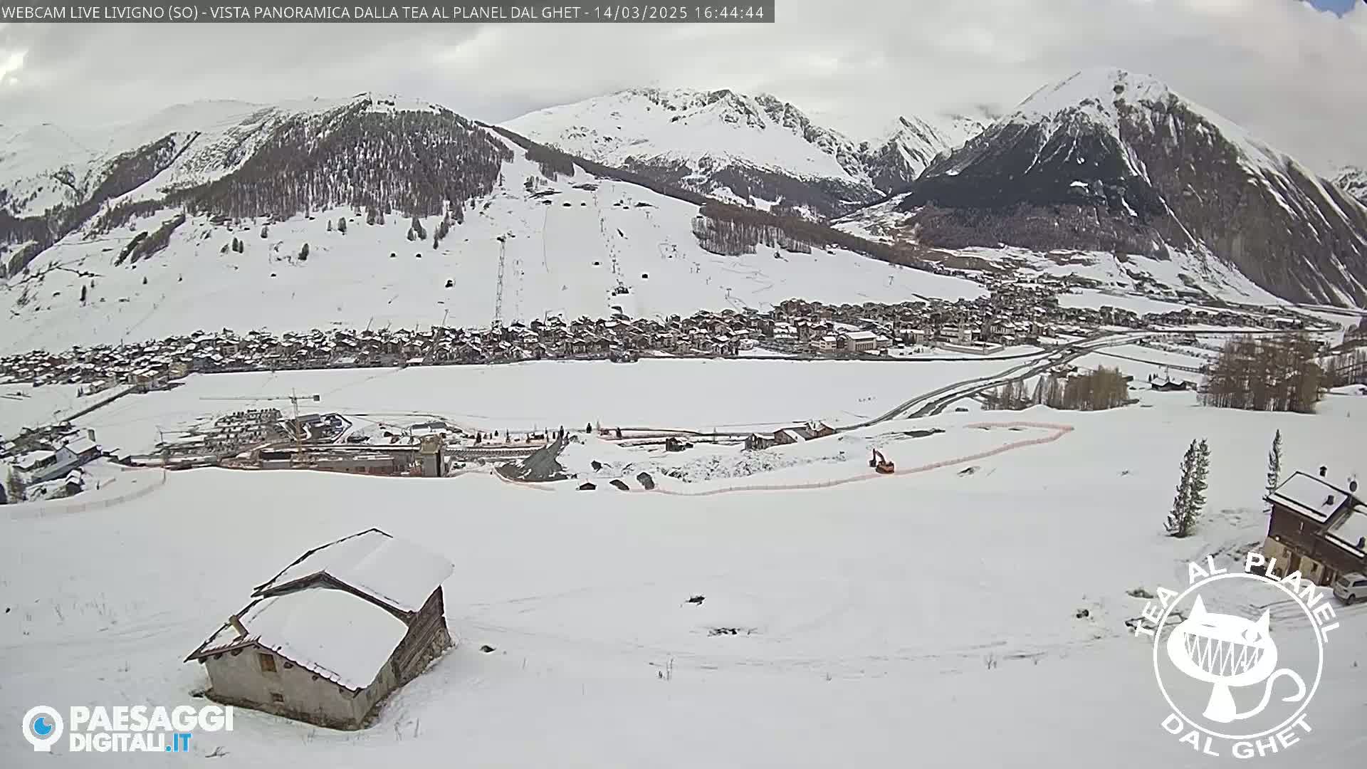 A snow-covered mountain village is seen from a high vantage point, with overcast skies and construction equipment visible in the foreground.