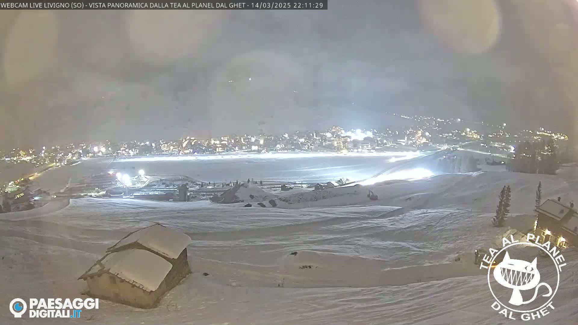 A snow-covered landscape at night shows a town illuminated in the distance under a hazy sky.