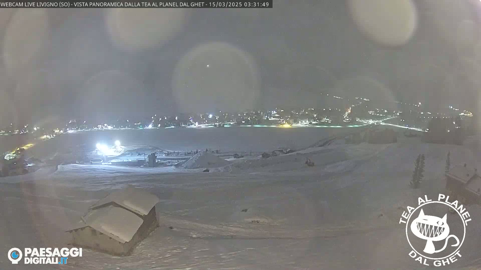 A nighttime, snowy panorama shows a snow-covered landscape with a small building in the foreground and a distant town illuminated with lights.
