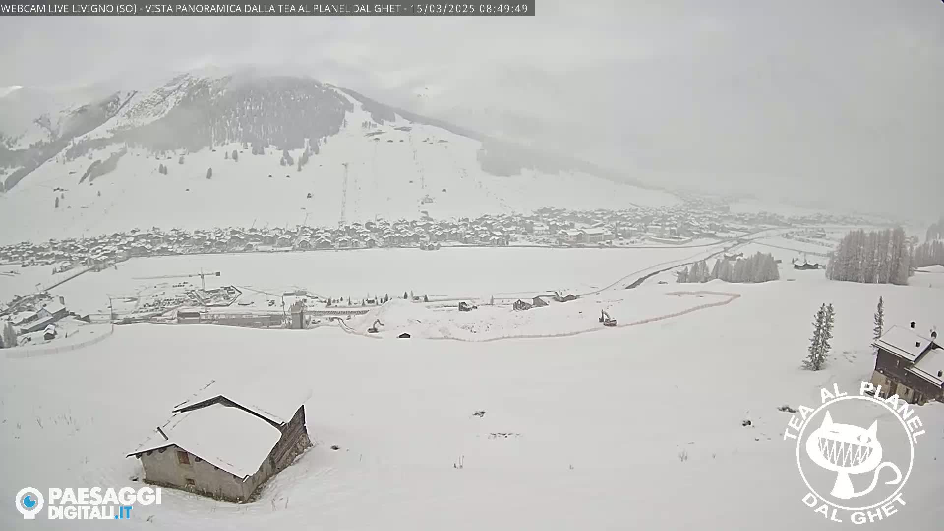 A snow-covered landscape shows a village in the valley below a snow-dusted mountain, with a small building in the foreground during a light snow.