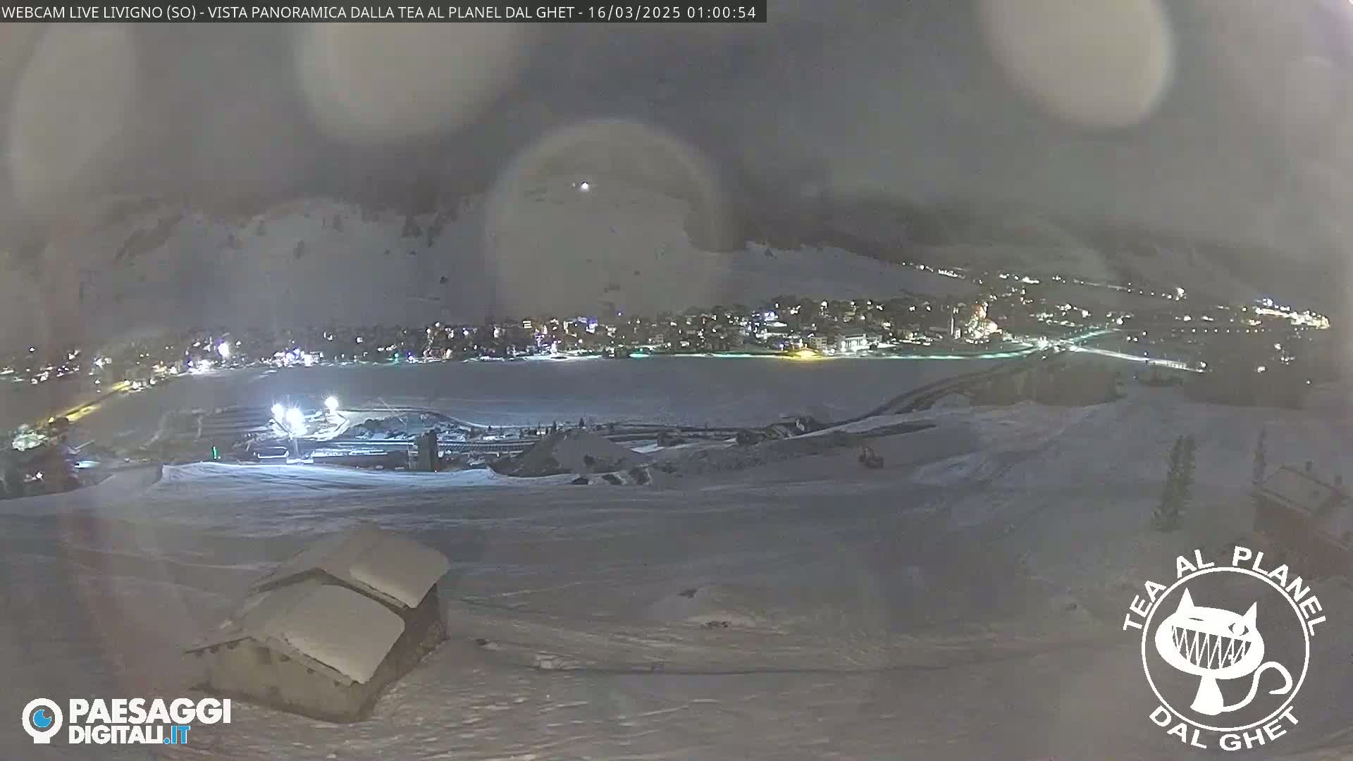 A nighttime, snowy scene shows a town illuminated below a hilltop, with a small snow-covered structure in the foreground.