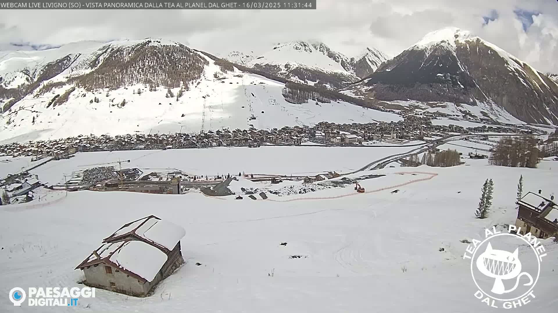 A snow-covered mountain village is viewed from a high vantage point on a partly cloudy day, with a small snow-covered building in the foreground.