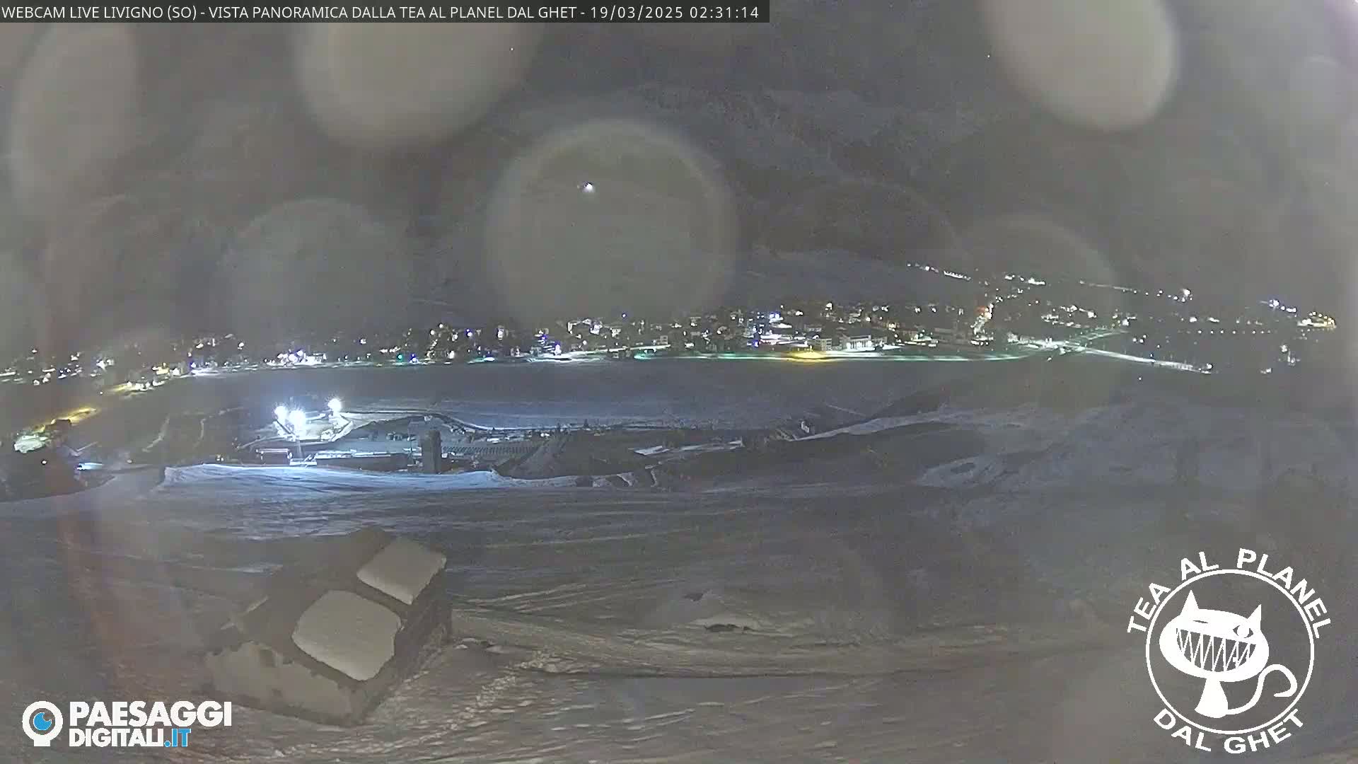 A nighttime, snowy landscape shows a town in the distance illuminated with lights under a dark, hazy sky.