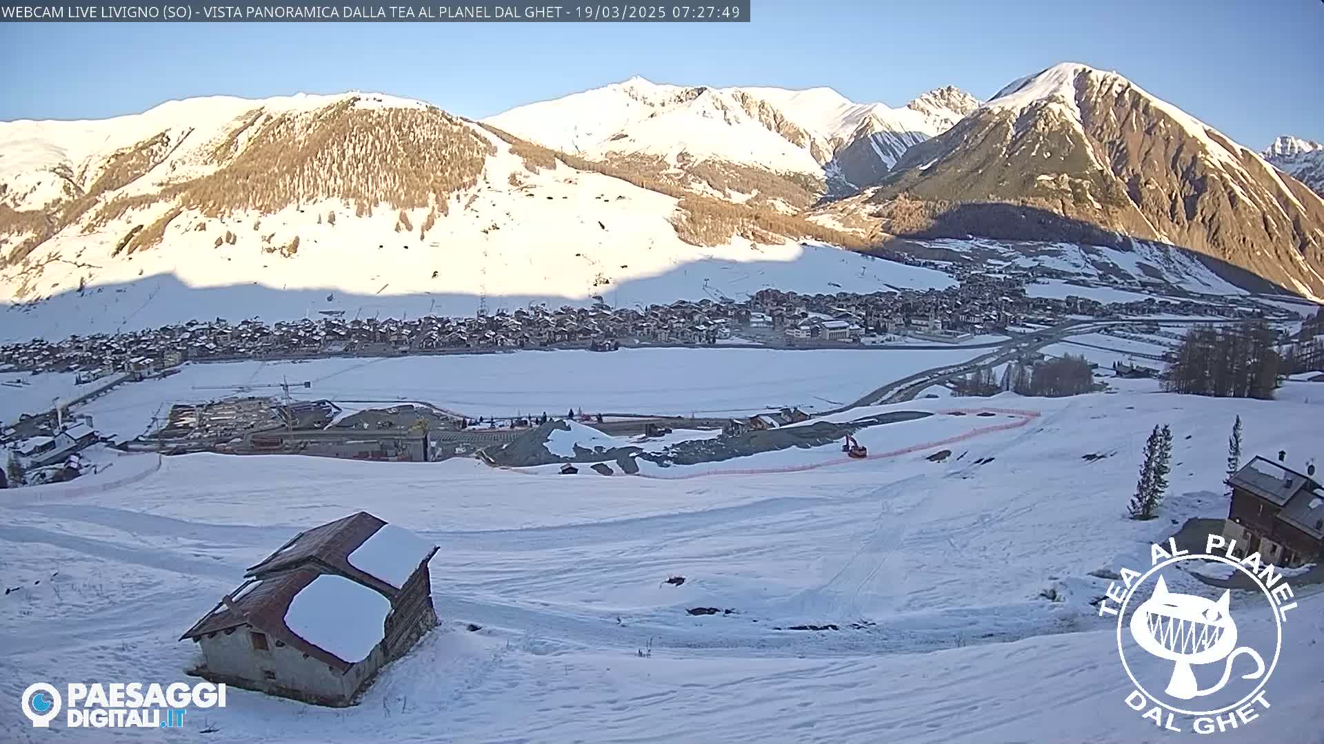 A snow-covered landscape shows a small village nestled in a valley between snow-capped mountains under a clear, bright sky.