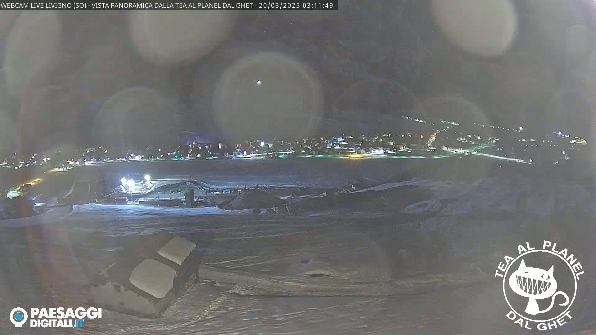 A nighttime, snowy landscape shows a distant town illuminated with lights under a dark sky with a faint halo around a light source.