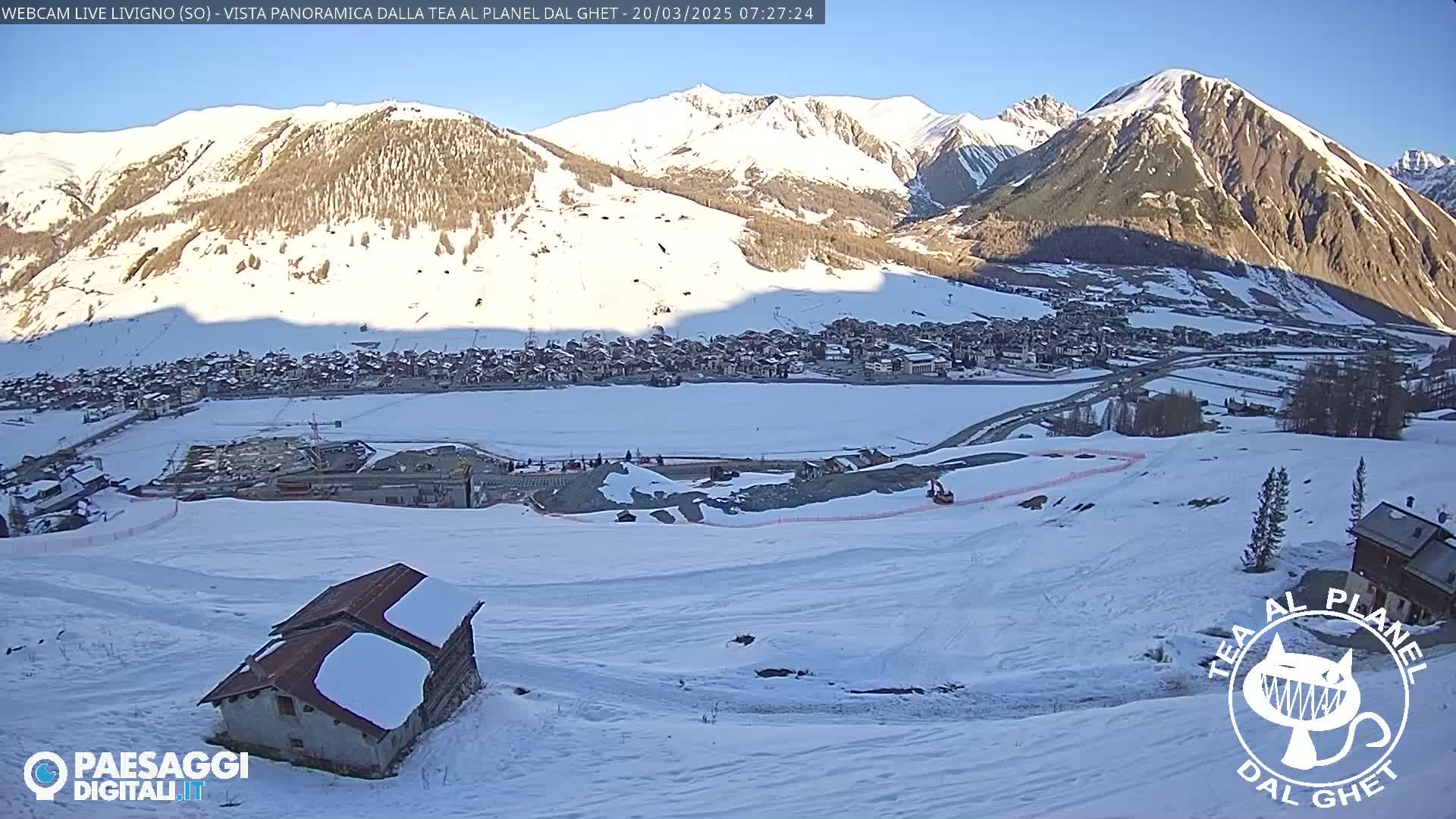 A snow-covered mountain village is nestled in a valley under a clear, sunny sky, with a small, snow-dusted building in the foreground.