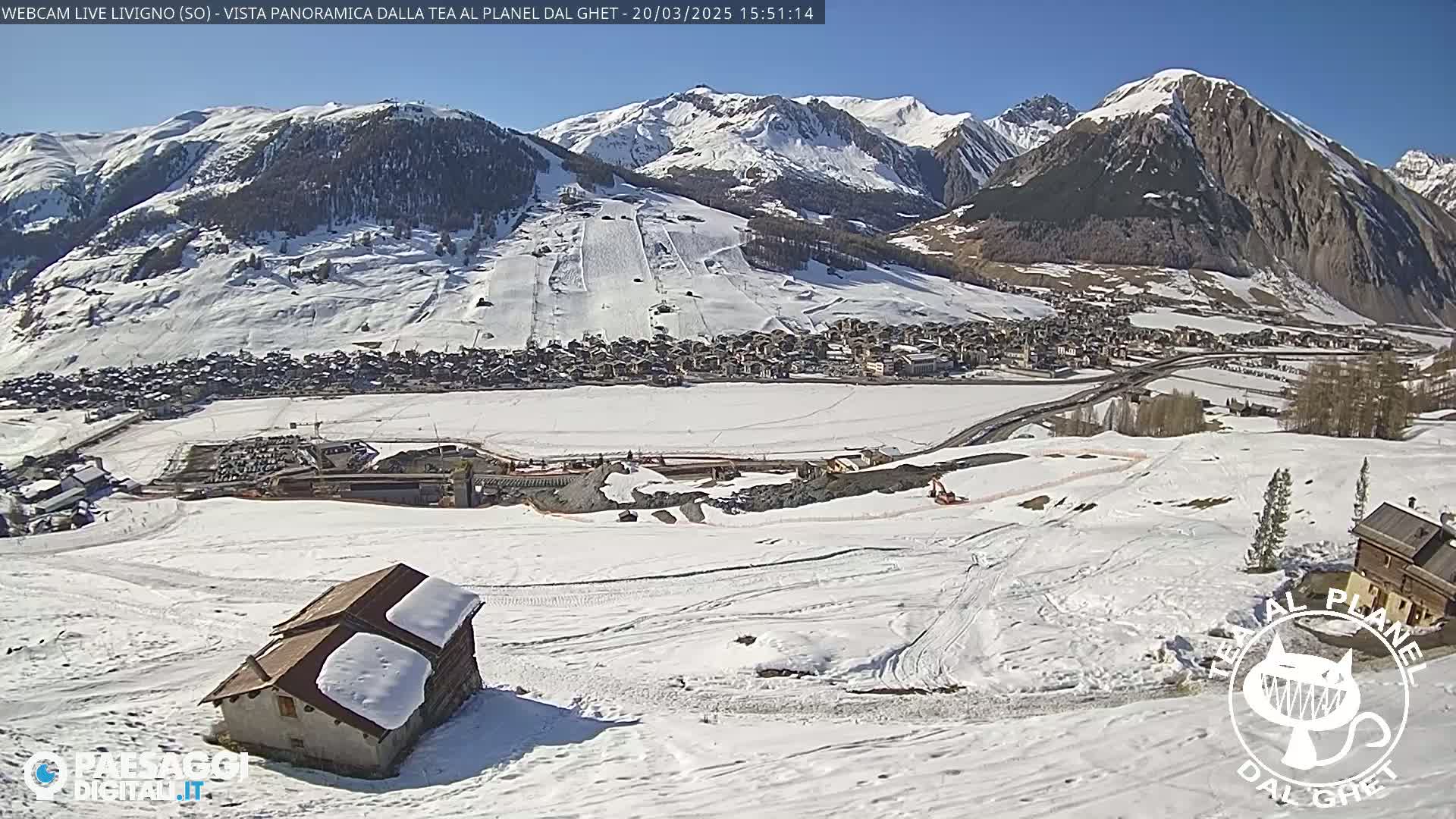 A snow-covered mountain valley town is seen from a high vantage point on a sunny day, with a small, snow-covered building in the foreground.
