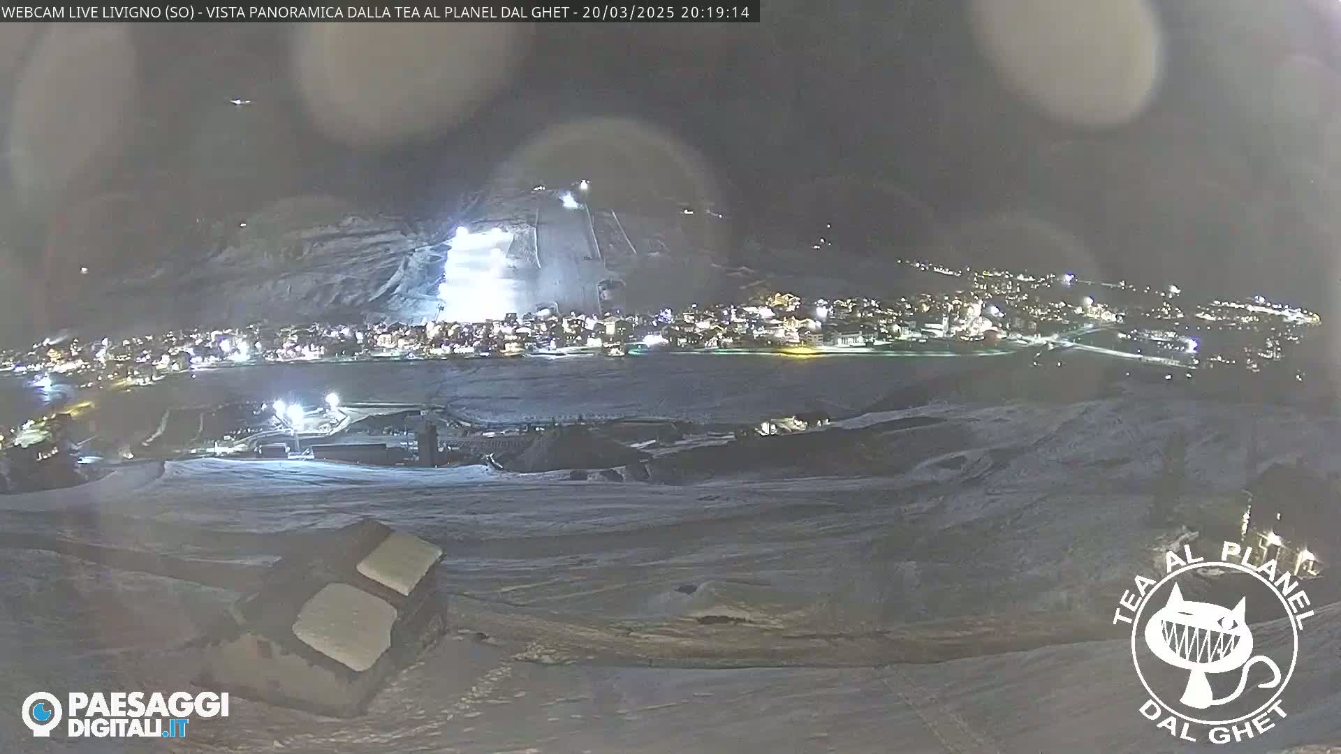 A nighttime, snow-covered landscape shows a town illuminated below, with ski slopes lit up in the background under a dark, possibly cloudy sky.