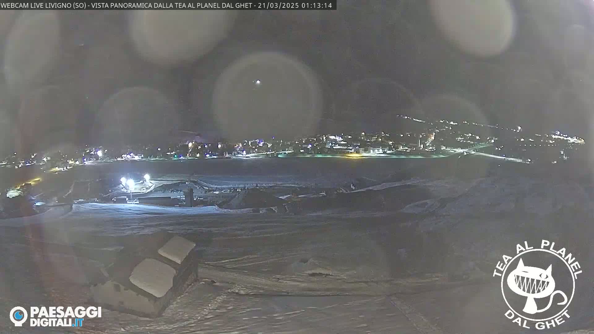 A nighttime, snowy landscape shows a distant town illuminated under a hazy sky.