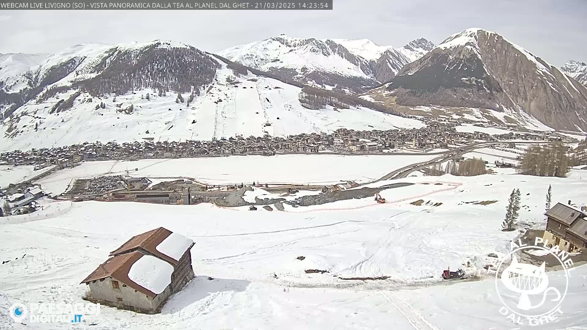 A snow-covered mountain village is seen from a high vantage point on a partly cloudy day, with a small building in the foreground and construction activity visible in the mid-ground.