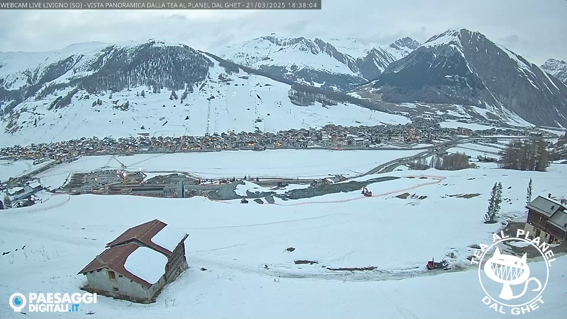 A snow-covered mountain village is visible below a snow-capped mountain range under an overcast sky.