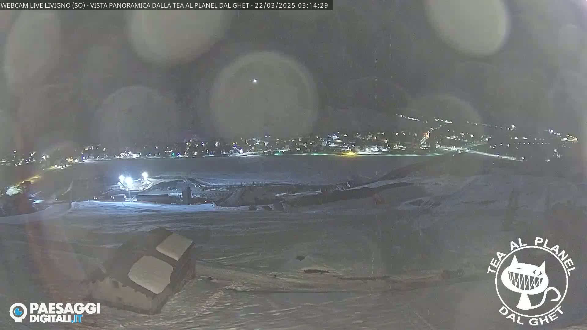 A nighttime, snowy panoramic view of a town illuminated by lights, with a structure in the foreground and a hazy sky.