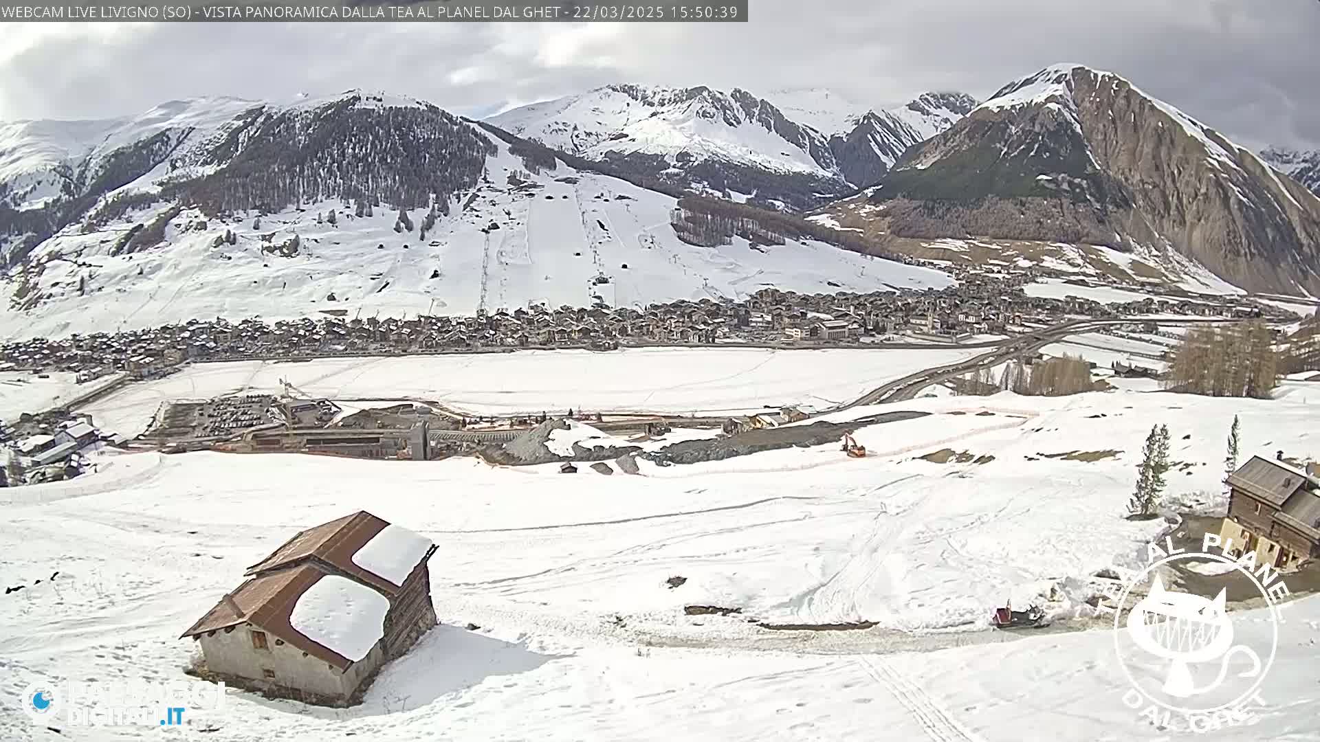 A snow-covered mountain valley town is viewed from a high vantage point on a partly cloudy day, with a small building partially buried in the snow in the foreground.