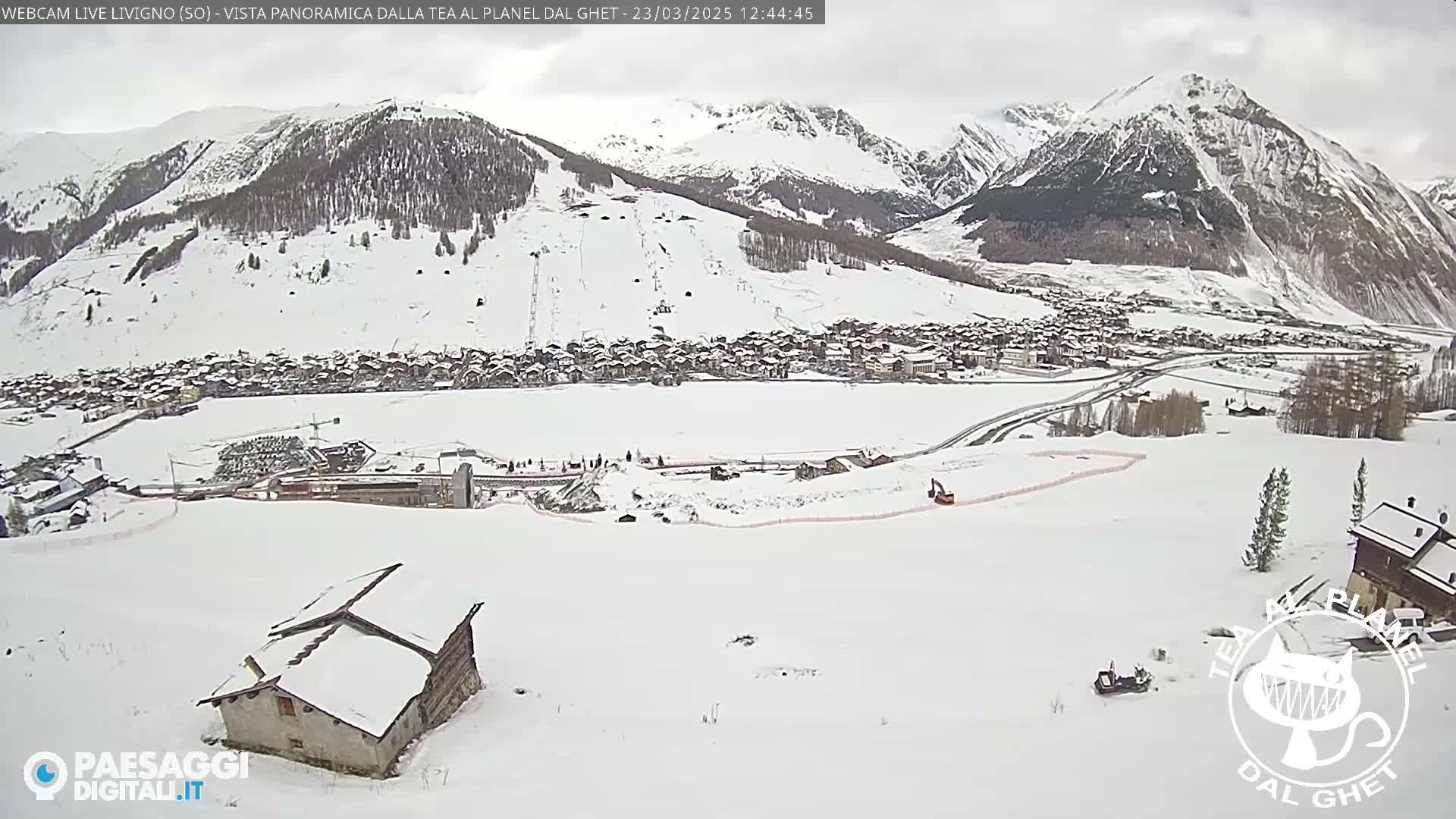 A snow-covered mountain village is visible from a high vantage point on a cloudy day, with snow-covered slopes and mountains in the background.