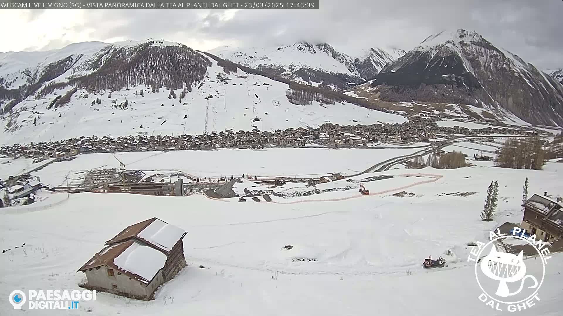 A snow-covered mountain village is nestled in a valley beneath snow-capped mountains under an overcast sky.