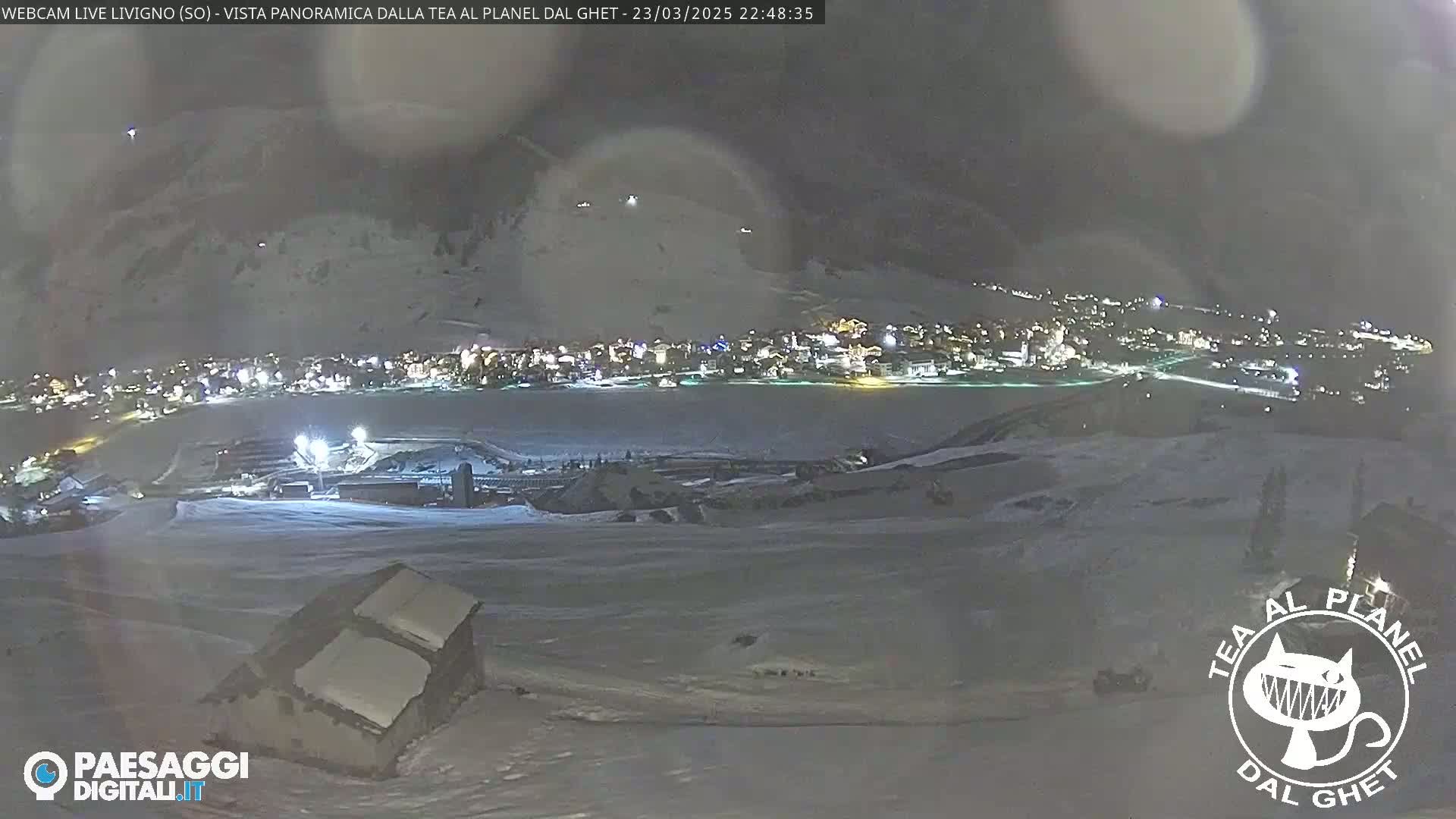 A nighttime, snowy landscape overlooks a town illuminated with lights, under a cloudy sky.