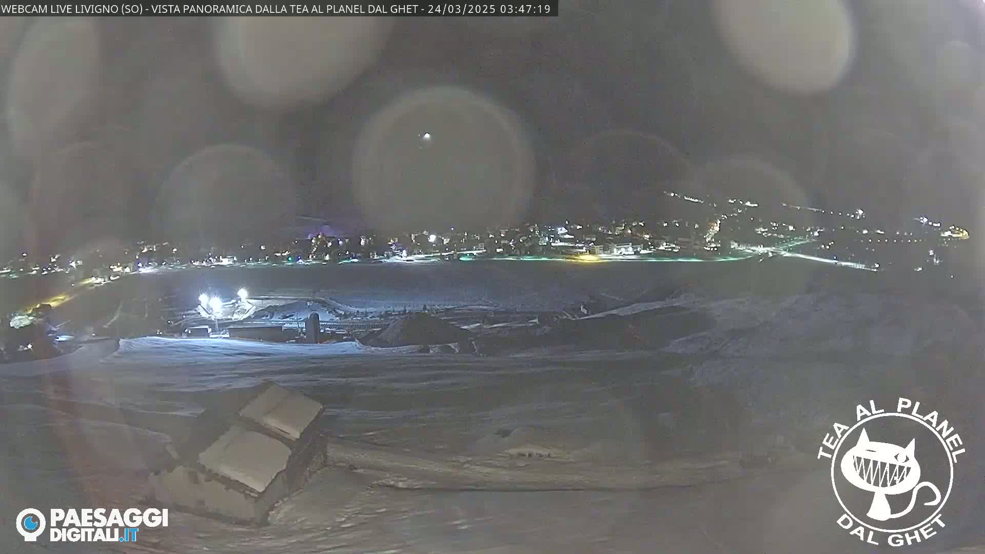A nighttime, snow-covered landscape shows a town in the distance, illuminated by lights, under a dark, hazy sky.