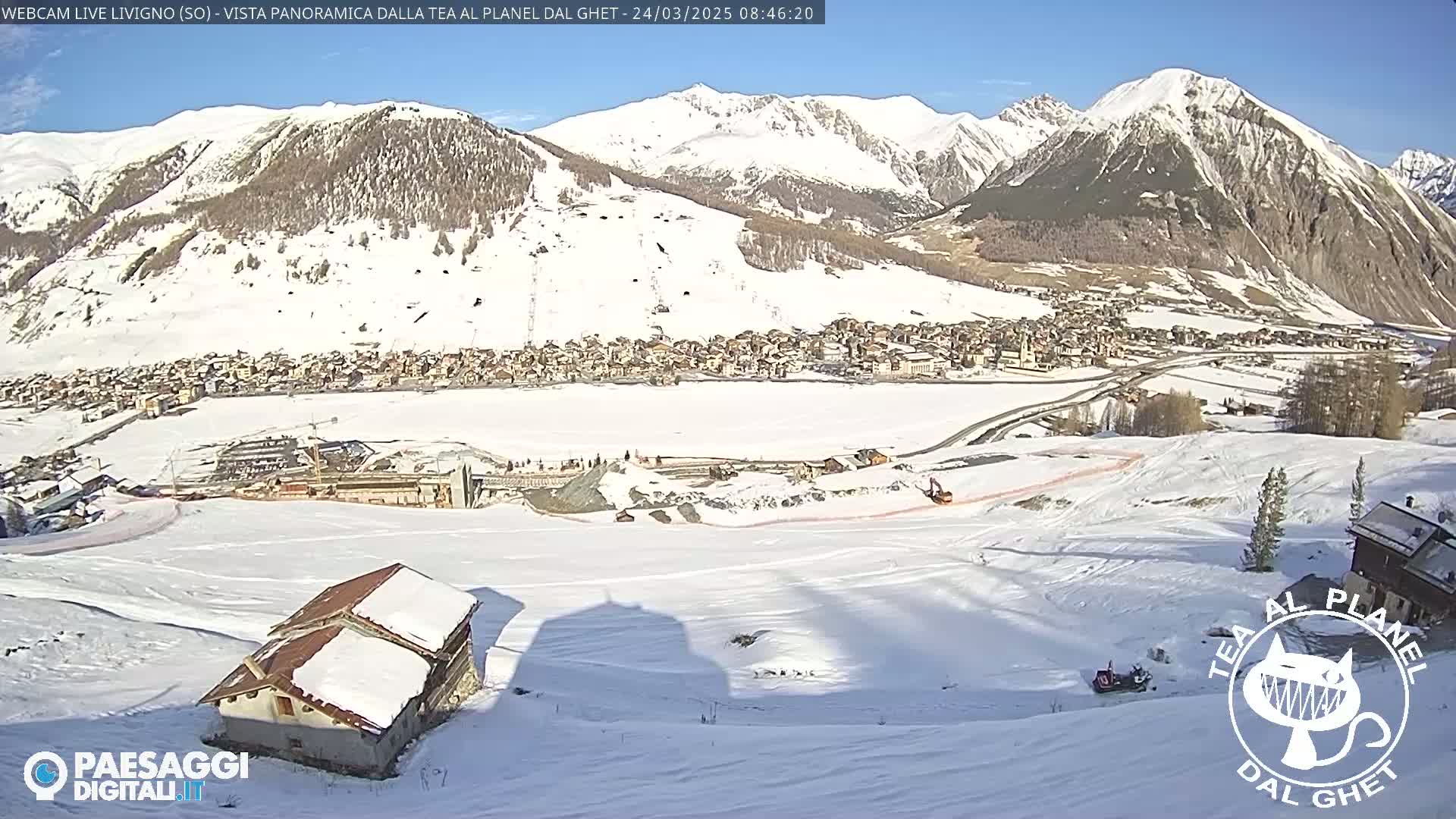 A snow-covered mountain village is seen from a high vantage point on a sunny day, with a small house and snowmobiles visible in the foreground.
