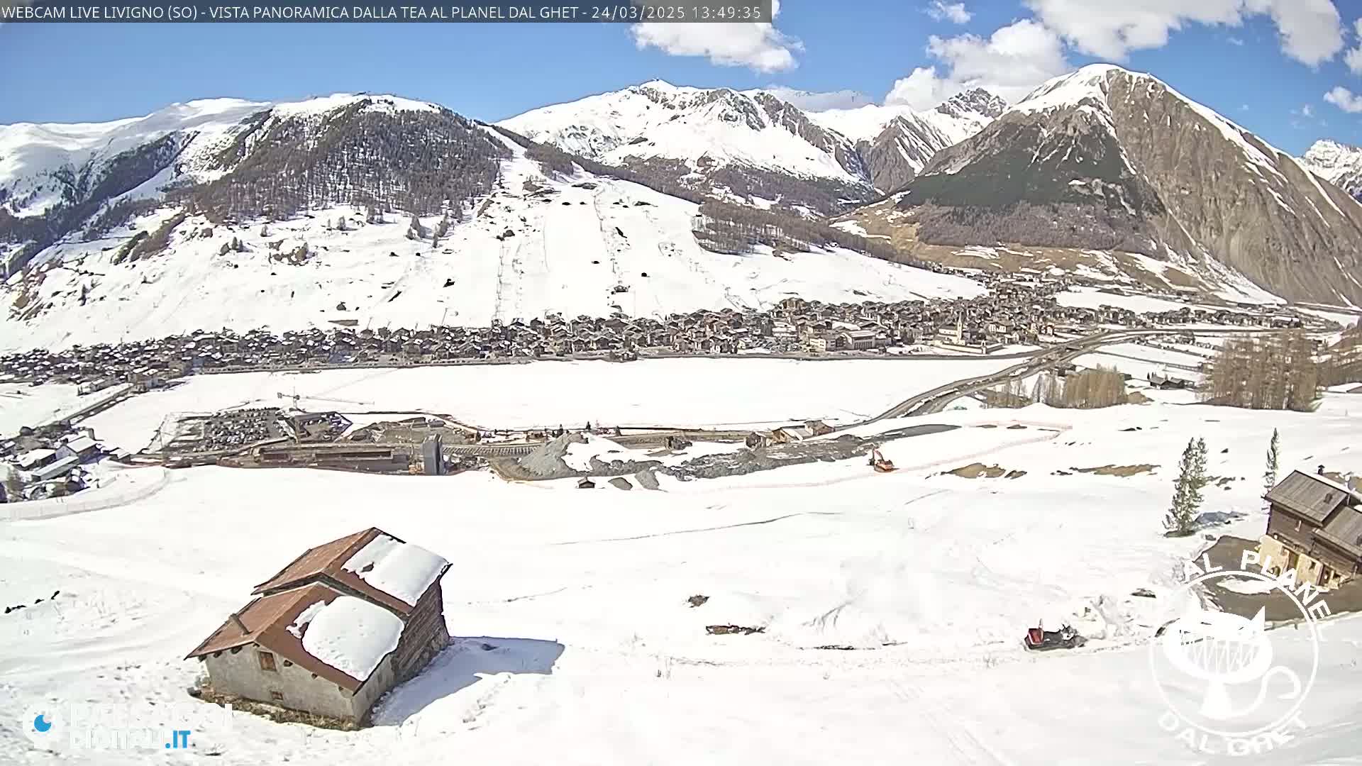 A snow-covered mountain village is viewed from a snowy hillside on a sunny day with some clouds.